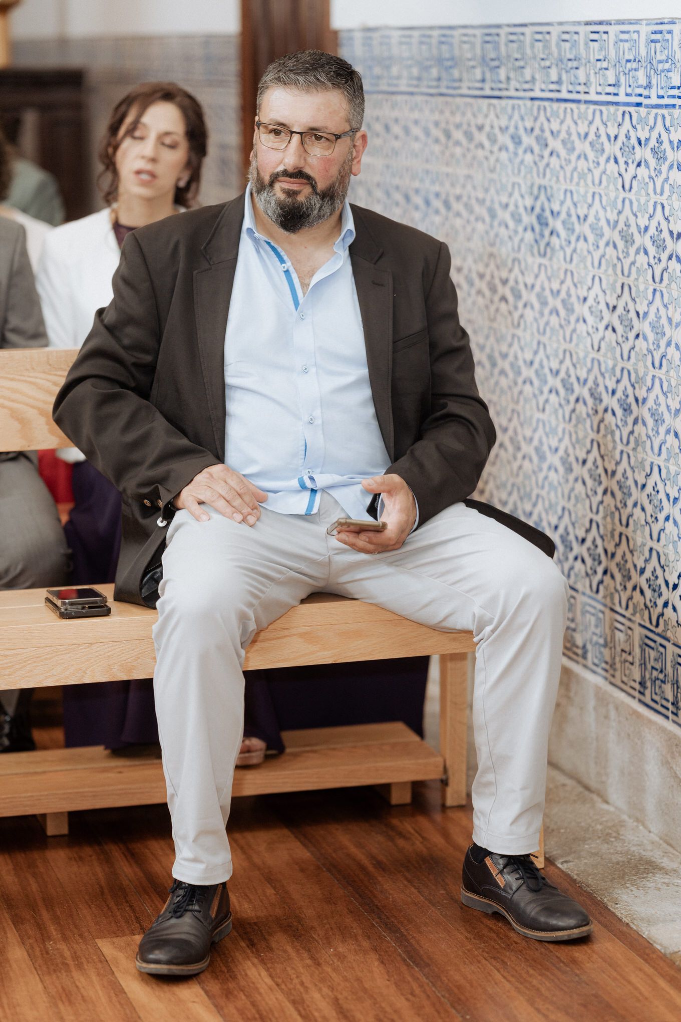 Professional sitting on wooden bench wearing brown blazer and light blue shirt in office with patterned tile wall background.