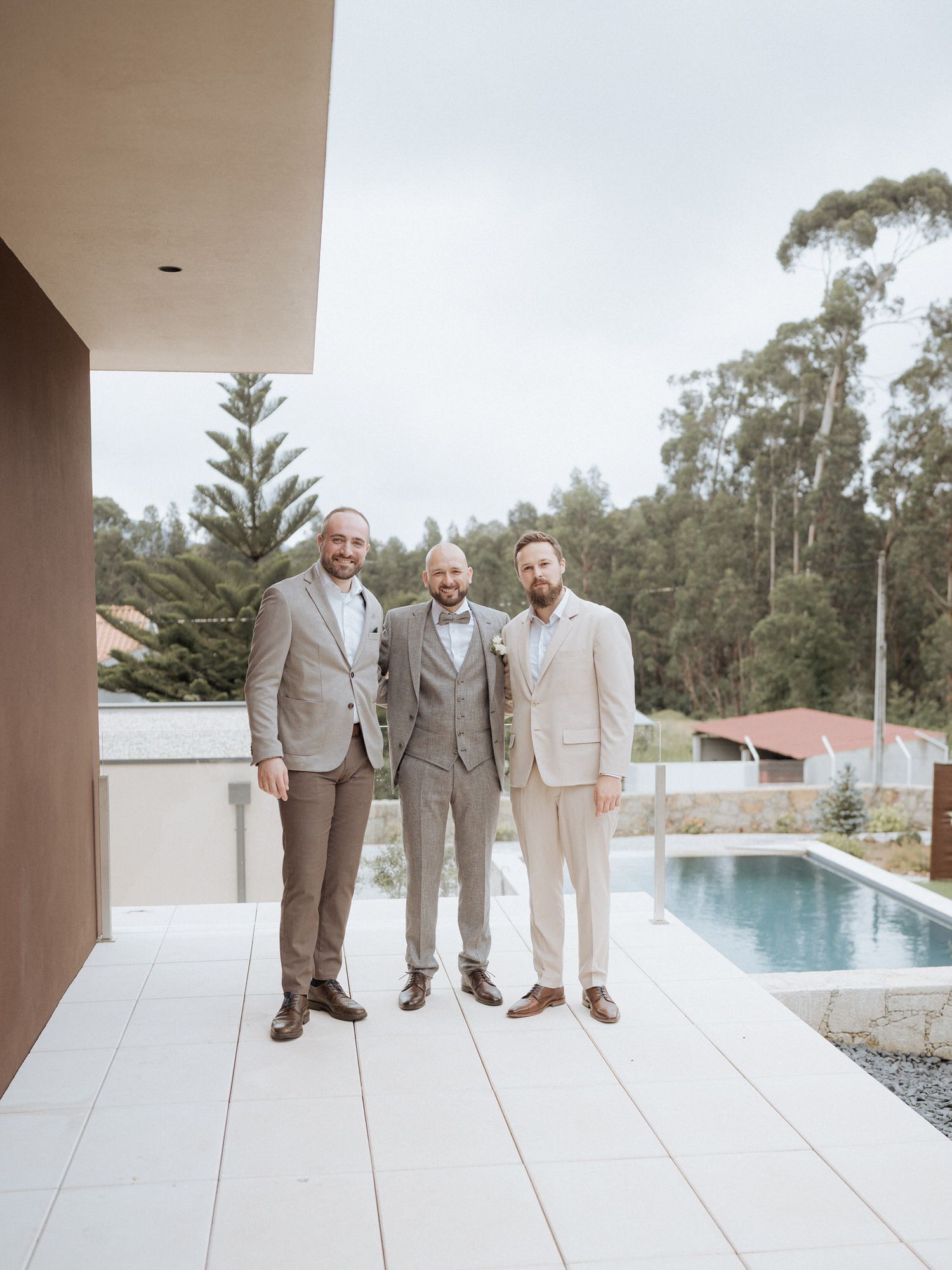 Three people in light colored suits stand together on a white tiled patio by a swimming pool with trees in background.