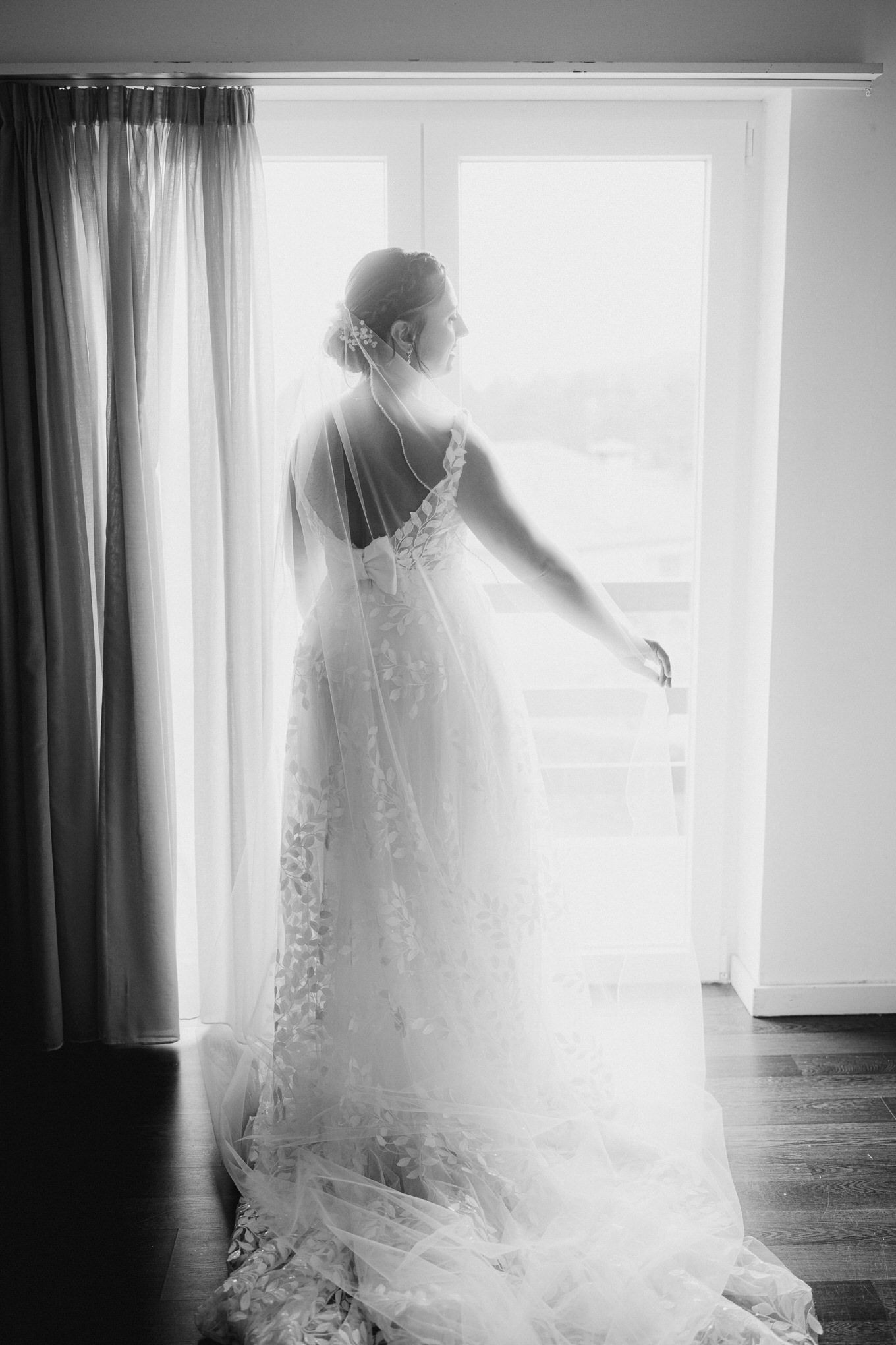 Black and white silhouette of a person in a lace wedding dress standing by a window basked in ethereal light.