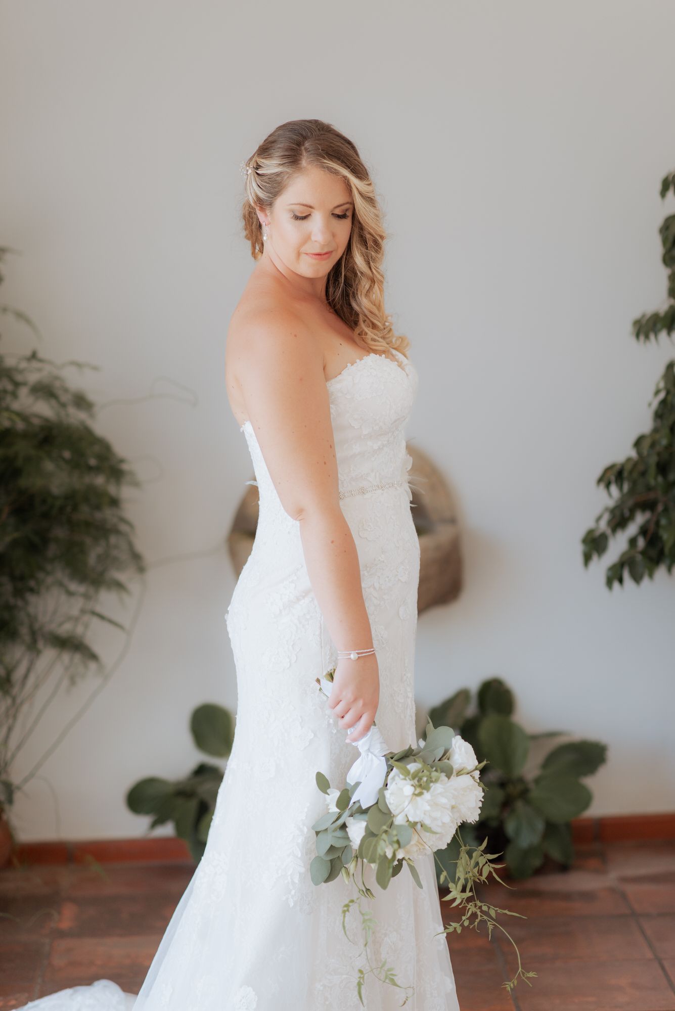 A bride in a strapless white lace wedding gown holds a delicate white and green floral bouquet while posing near indoor plants.