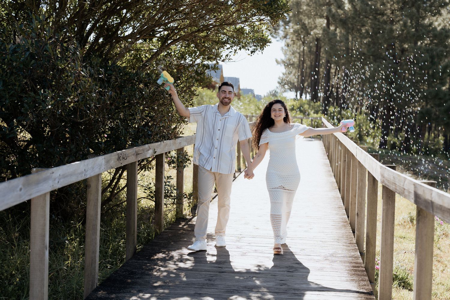 Duas pessoas caminham de mãos dadas por um calçadão de madeira ensolarado, cercado por vegetação.
