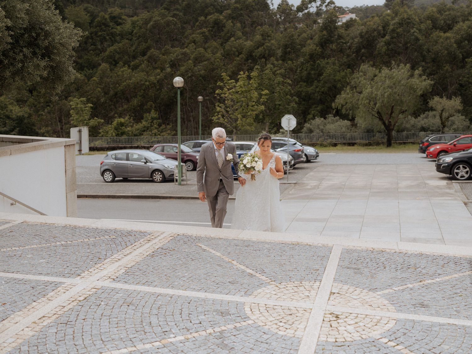 A wedding couple walks across a paved courtyard with a parking lot and trees in the background.