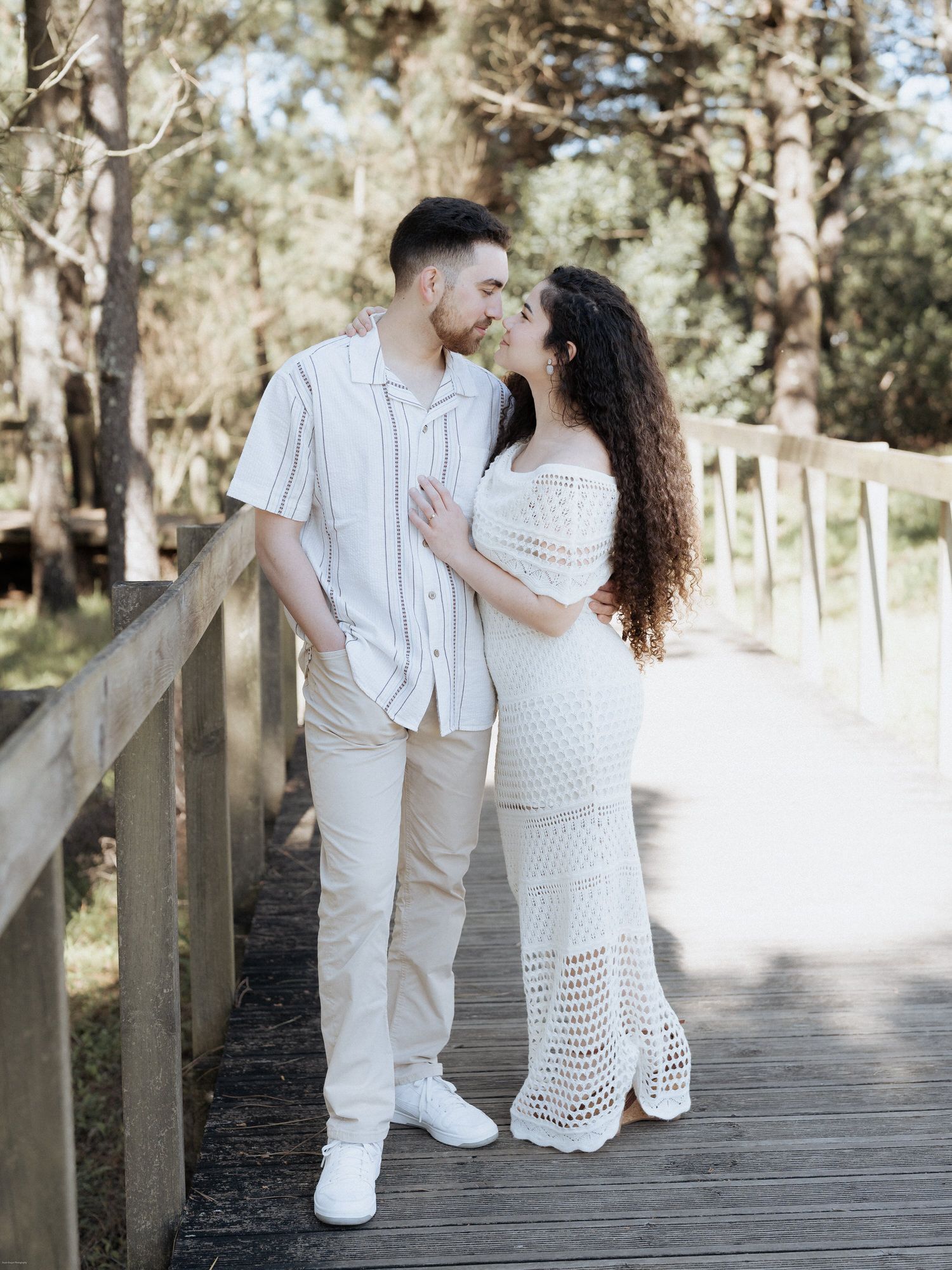 Um casal romântico que veste roupas brancas partilha um momento íntimo numa passarela de madeira num cenário natural.
