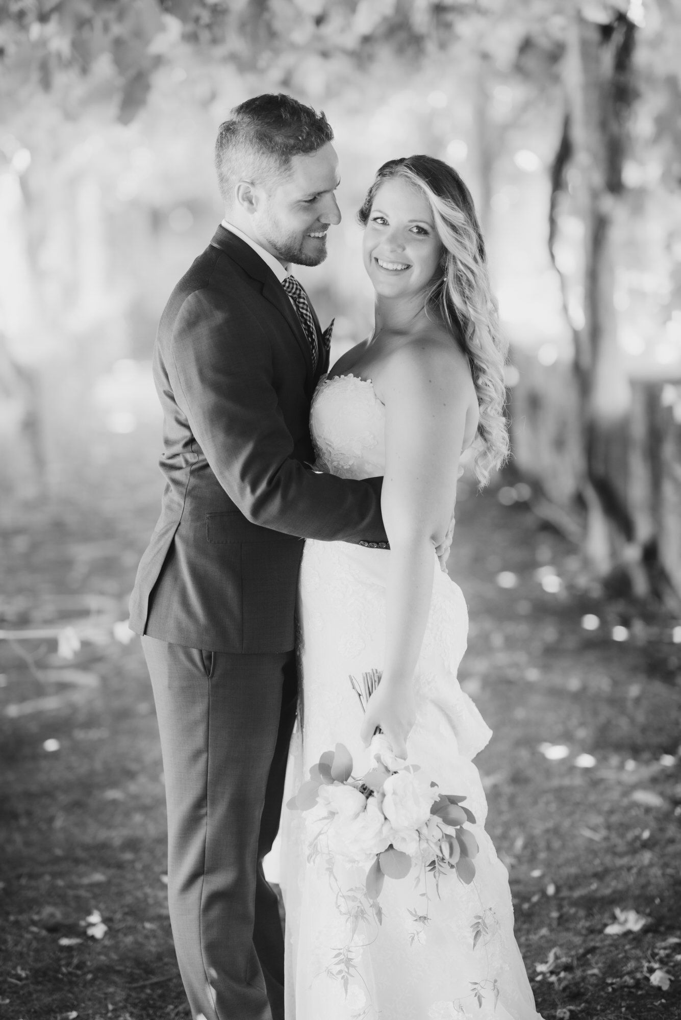 Black and white wedding photo of a couple embracing outdoors in a romantic woodland setting with the bride in a flowing gown.