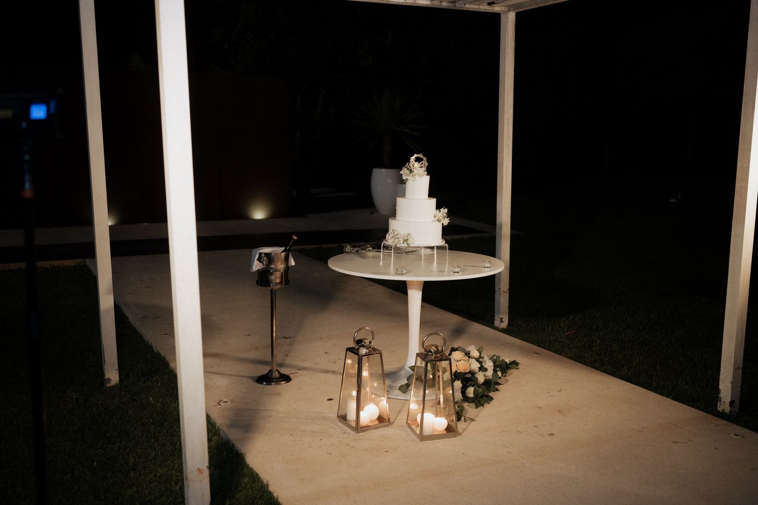 A white wedding cake displayed on a table outdoors at night with lanterns glowing beneath it.