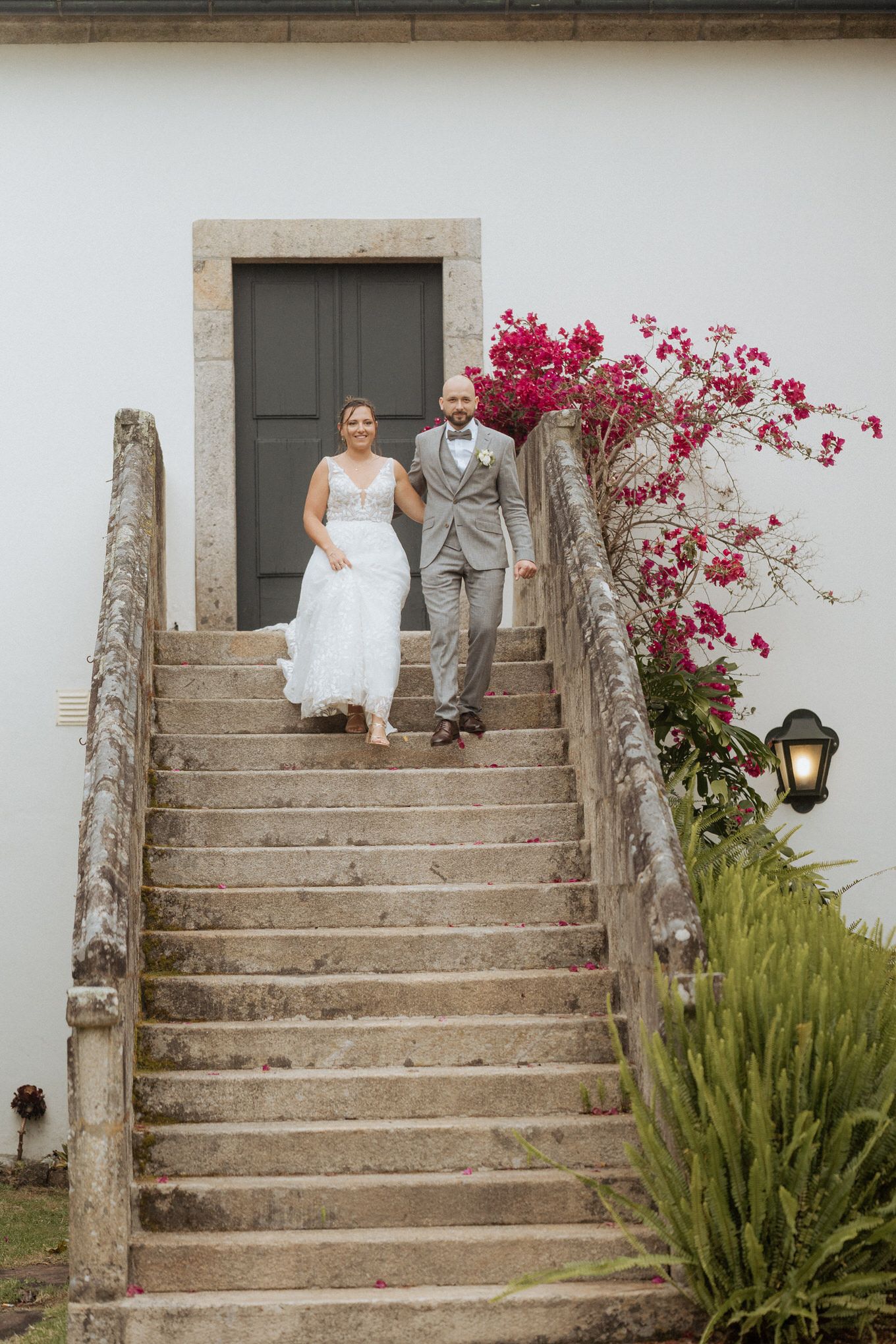 Two people descend stone steps at an elegant venue with pink bougainvillea flowers and a lantern beside an old doorway.
