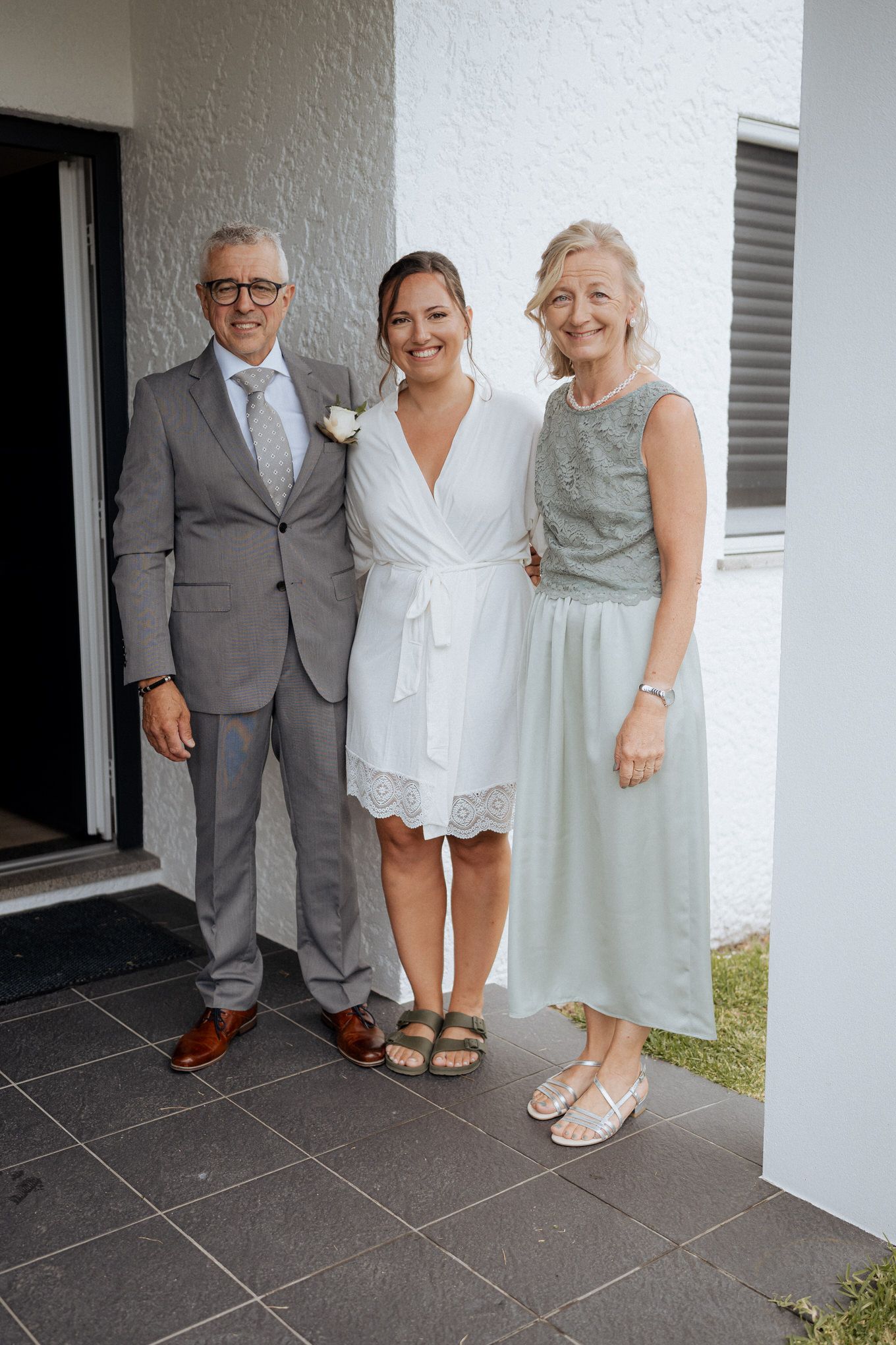 Three people in formal attire pose together on a tiled patio outside a white building.
