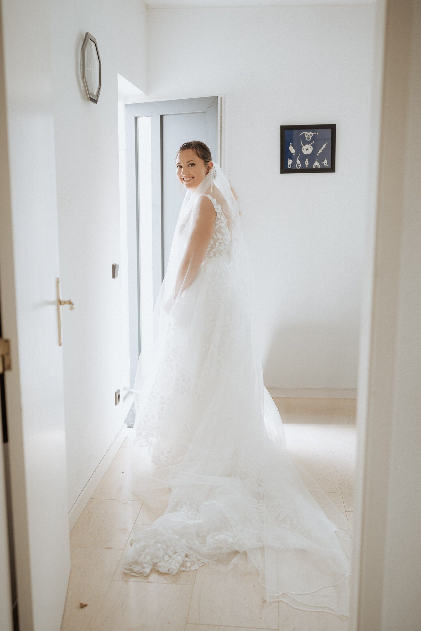 A bride in an elegant white lace wedding gown and long veil stands in a bright hallway near a doorway.