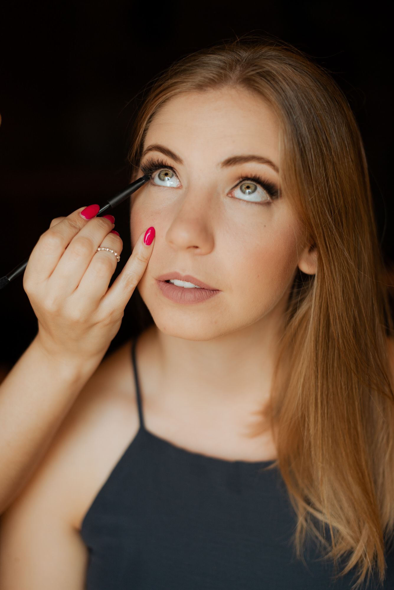 Makeup artist applying mascara to woman's eyelashes with pink nail polish in dark studio setting.