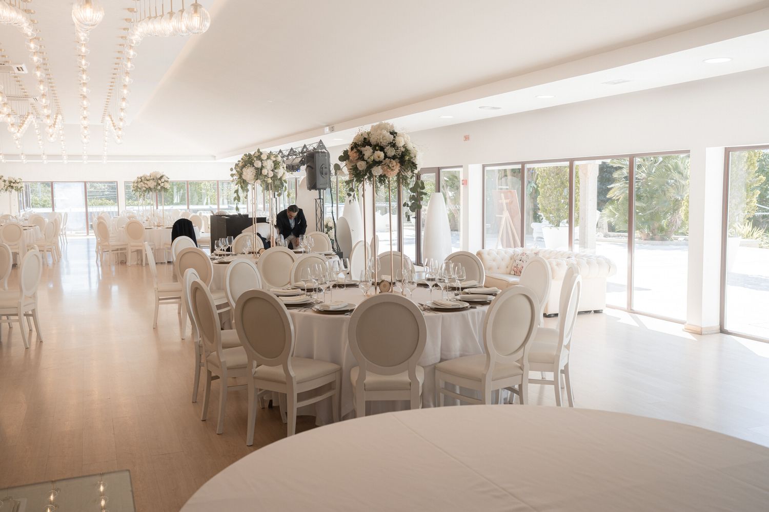 A bright white wedding venue with floor-to-ceiling windows, round dining tables, chandeliers and natural lighting.