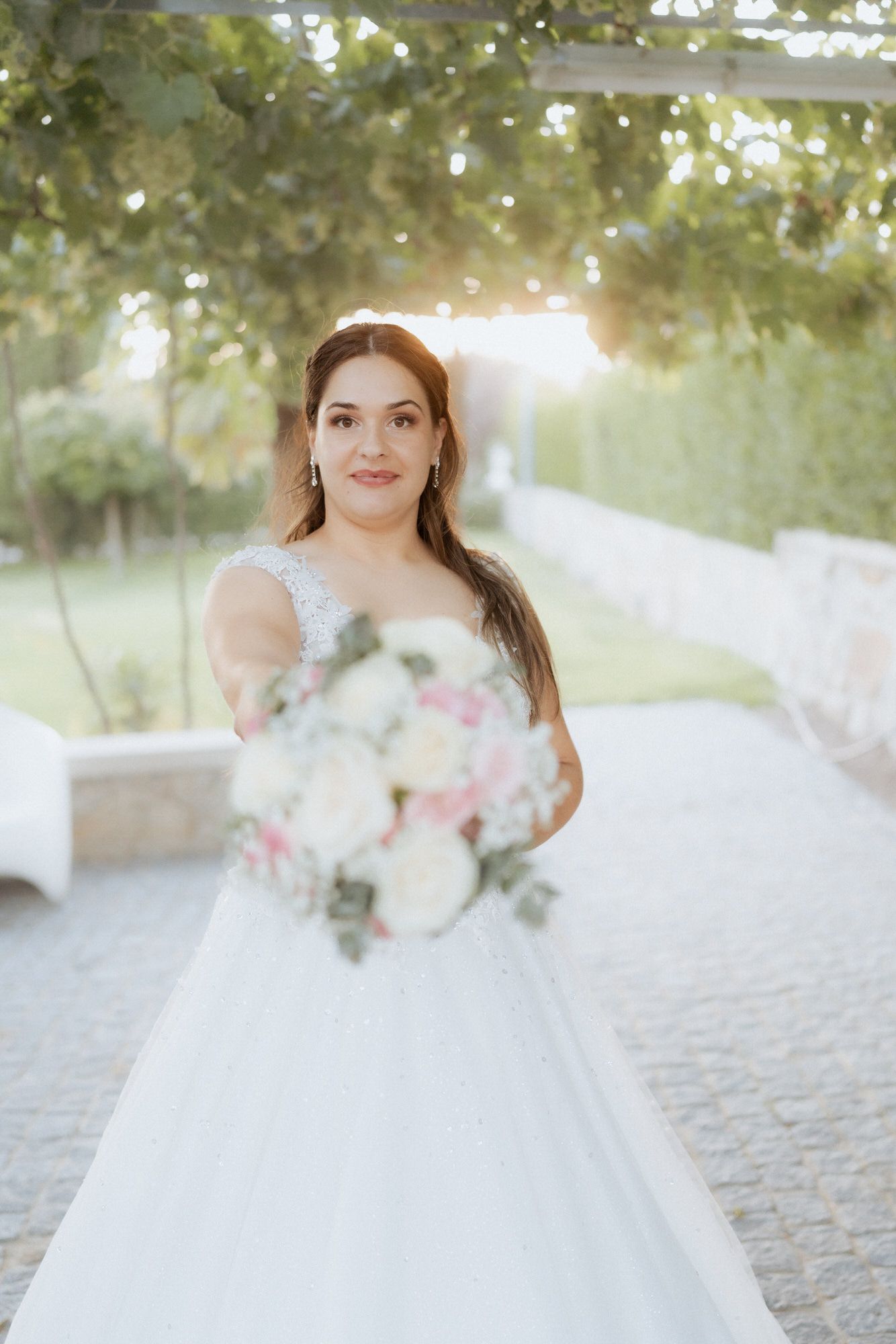 A bride in a flowing white ballgown wedding dress holds out a pastel bouquet of flowers in an outdoor garden setting.