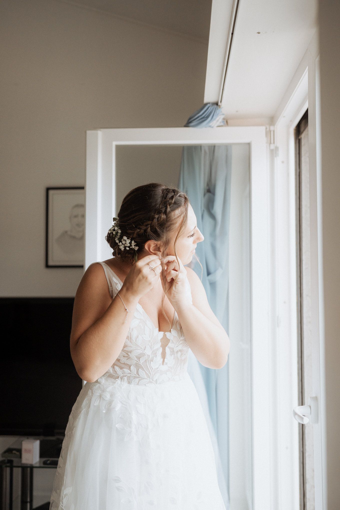 Person putting on jewelry while wearing an elegant white lace wedding dress near a window with soft natural light.