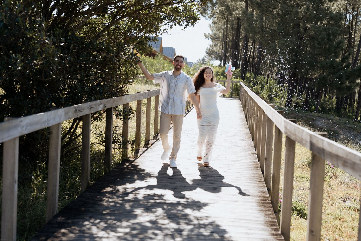 Um casal em trajes brancos de verão caminha de mãos dadas por um calçadão de madeira cercado por árvores em um dia ensolarado.