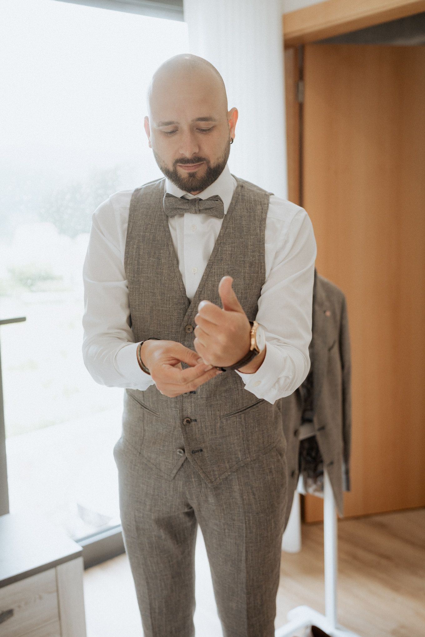 Well-dressed groom in grey tweed vest and bowtie adjusts his cufflinks while getting ready in a sunlit room.