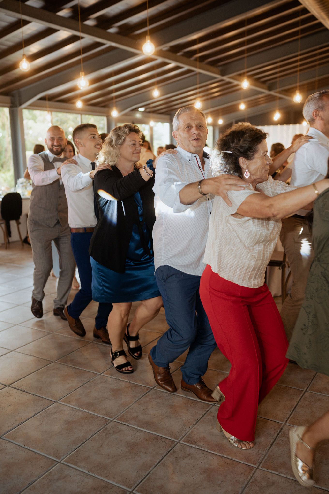 Joyful group of guests form a conga line dancing and celebrating at an indoor event with wooden beamed ceiling.
