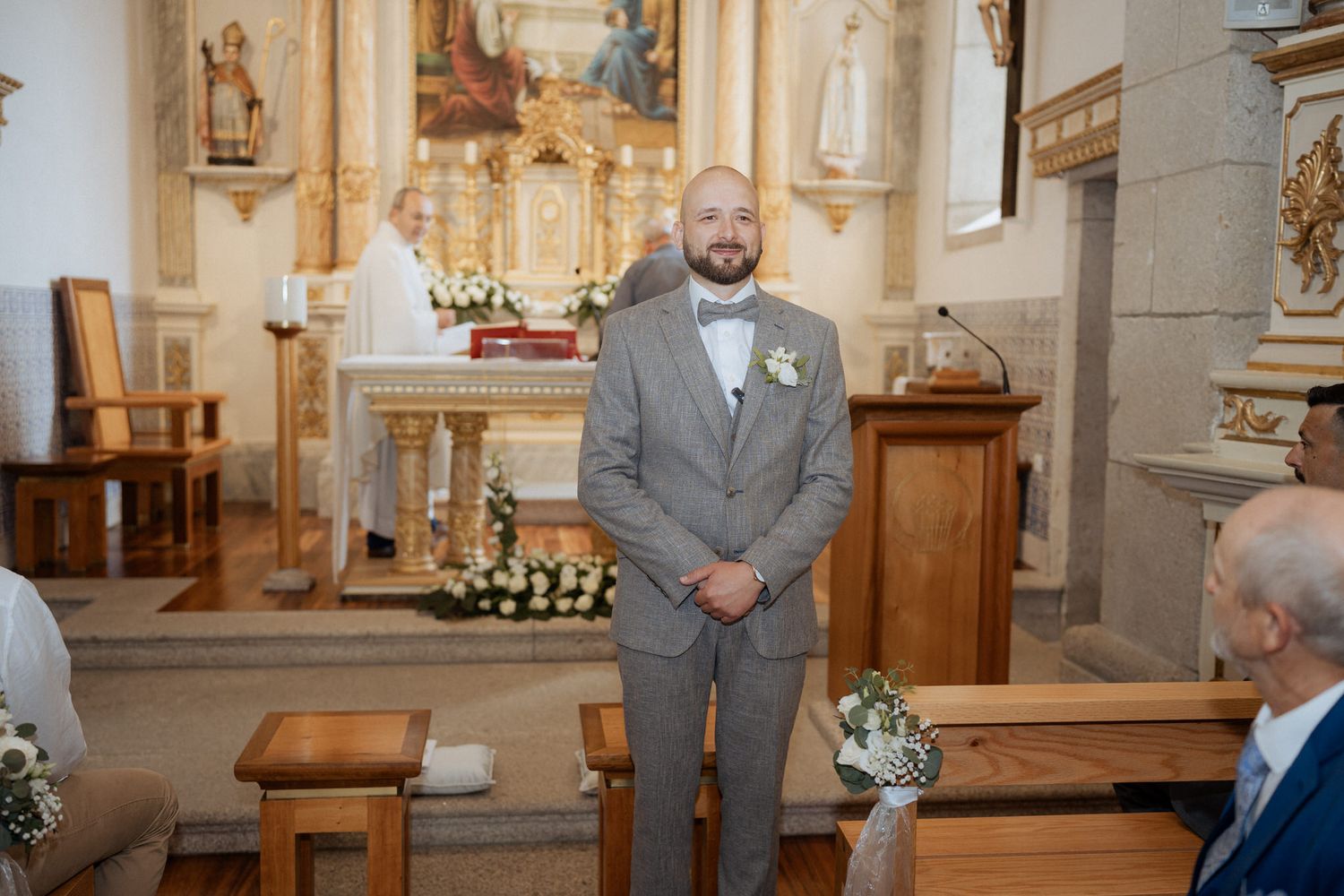 A groom in a light gray suit stands at the altar of a traditional church decorated with floral arrangements.
