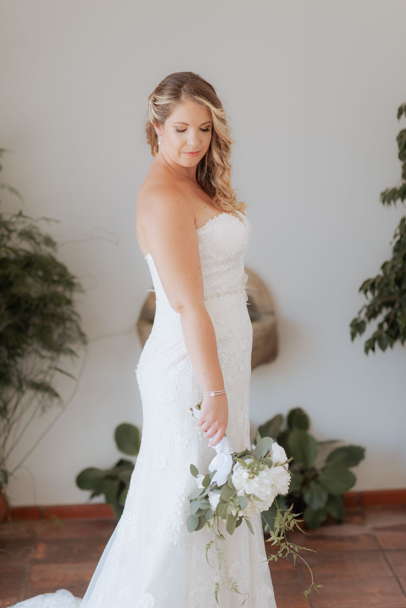 Elegant strapless lace wedding gown with flowing train and white floral bouquet against terracotta tile and greenery backdrop.