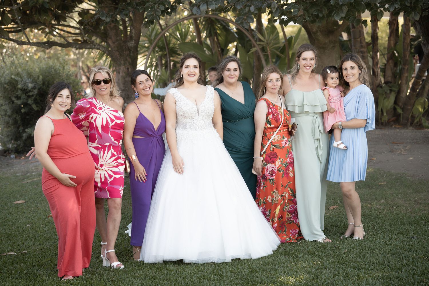 A group of women in colorful dresses and gowns pose together on a lawn with trees in the background.
