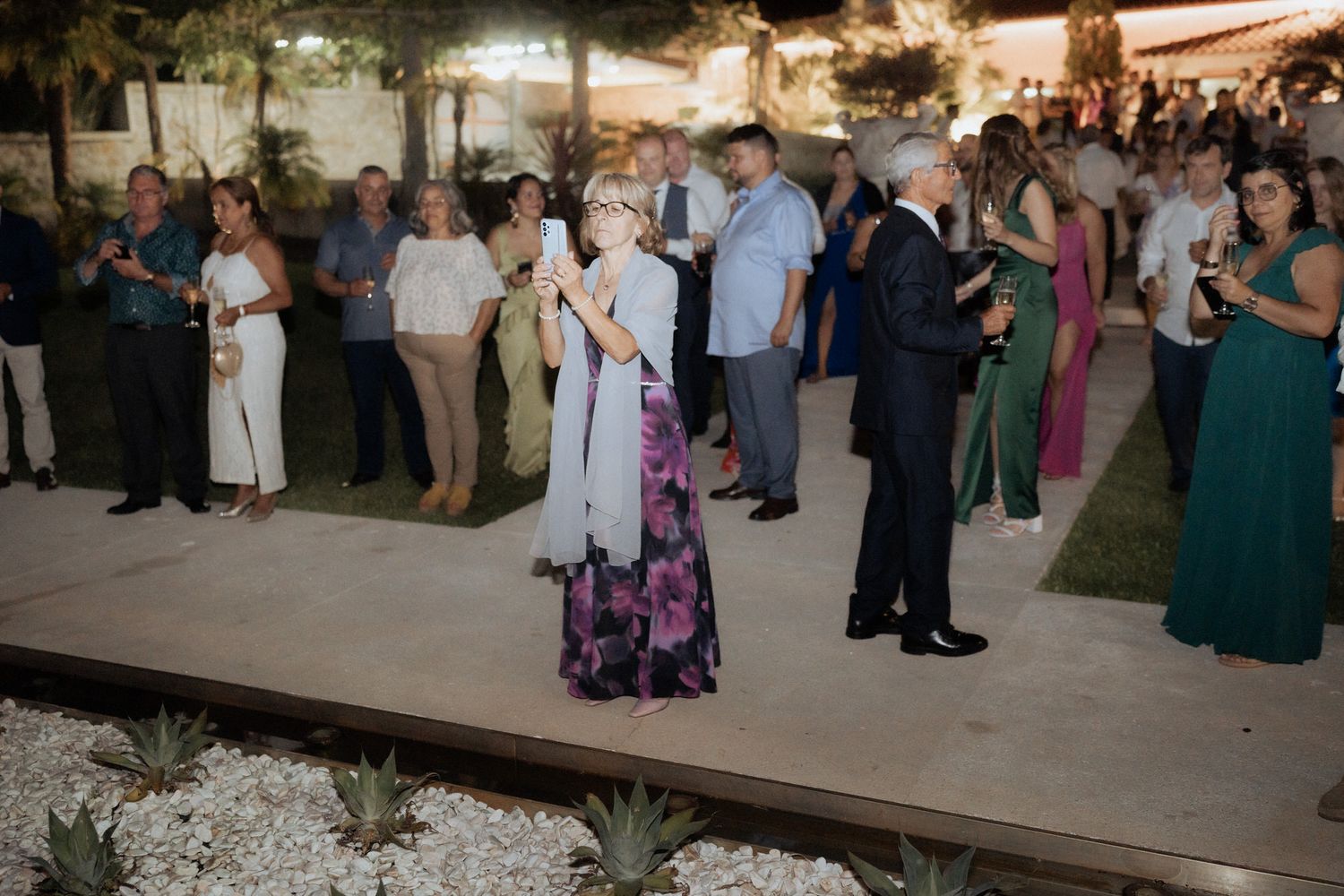 Wedding guests gather on an outdoor patio during an evening reception with decorative lighting and landscaping.