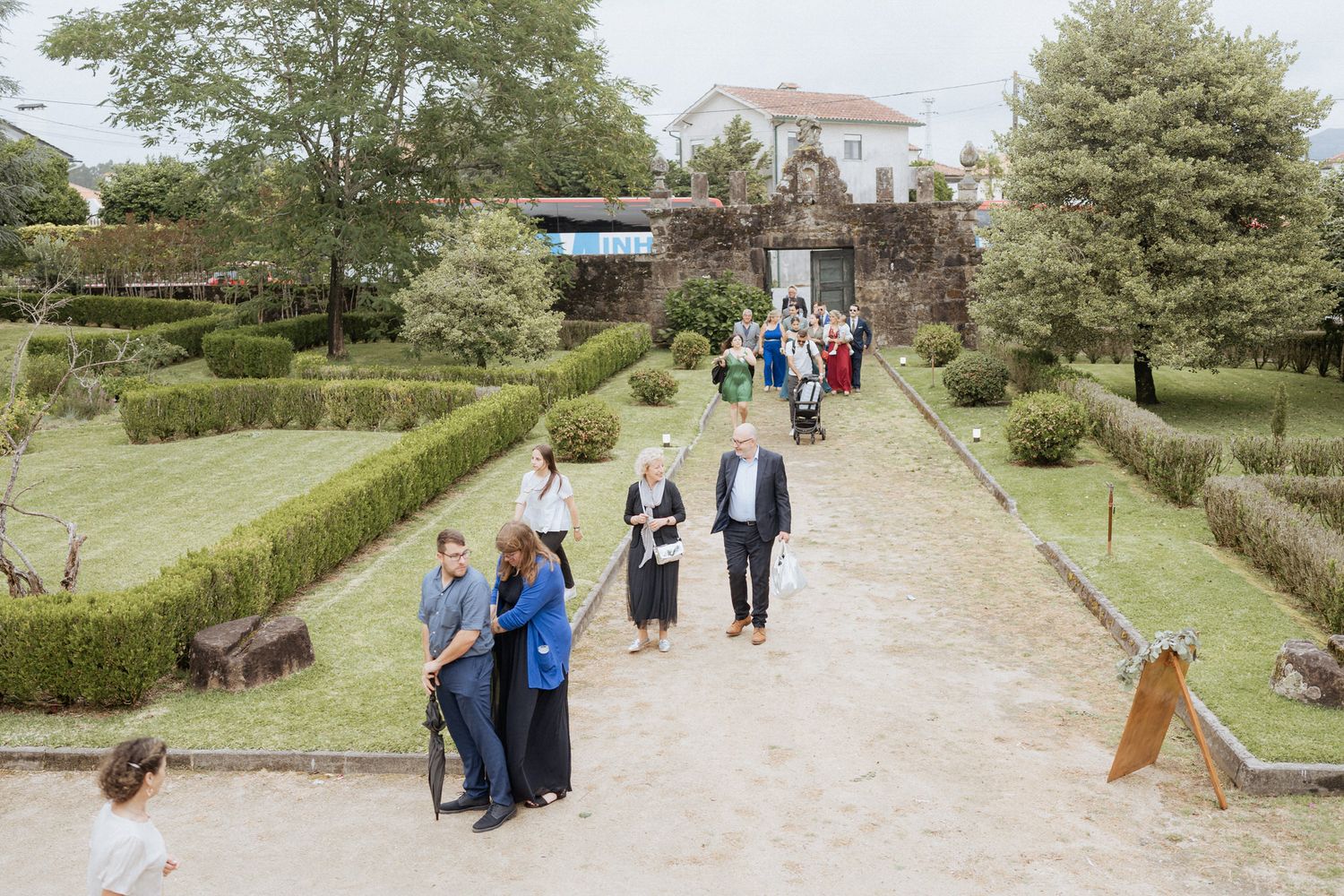 Visitors walk along a manicured garden path leading to a historic mansion with neatly trimmed hedges and trees.