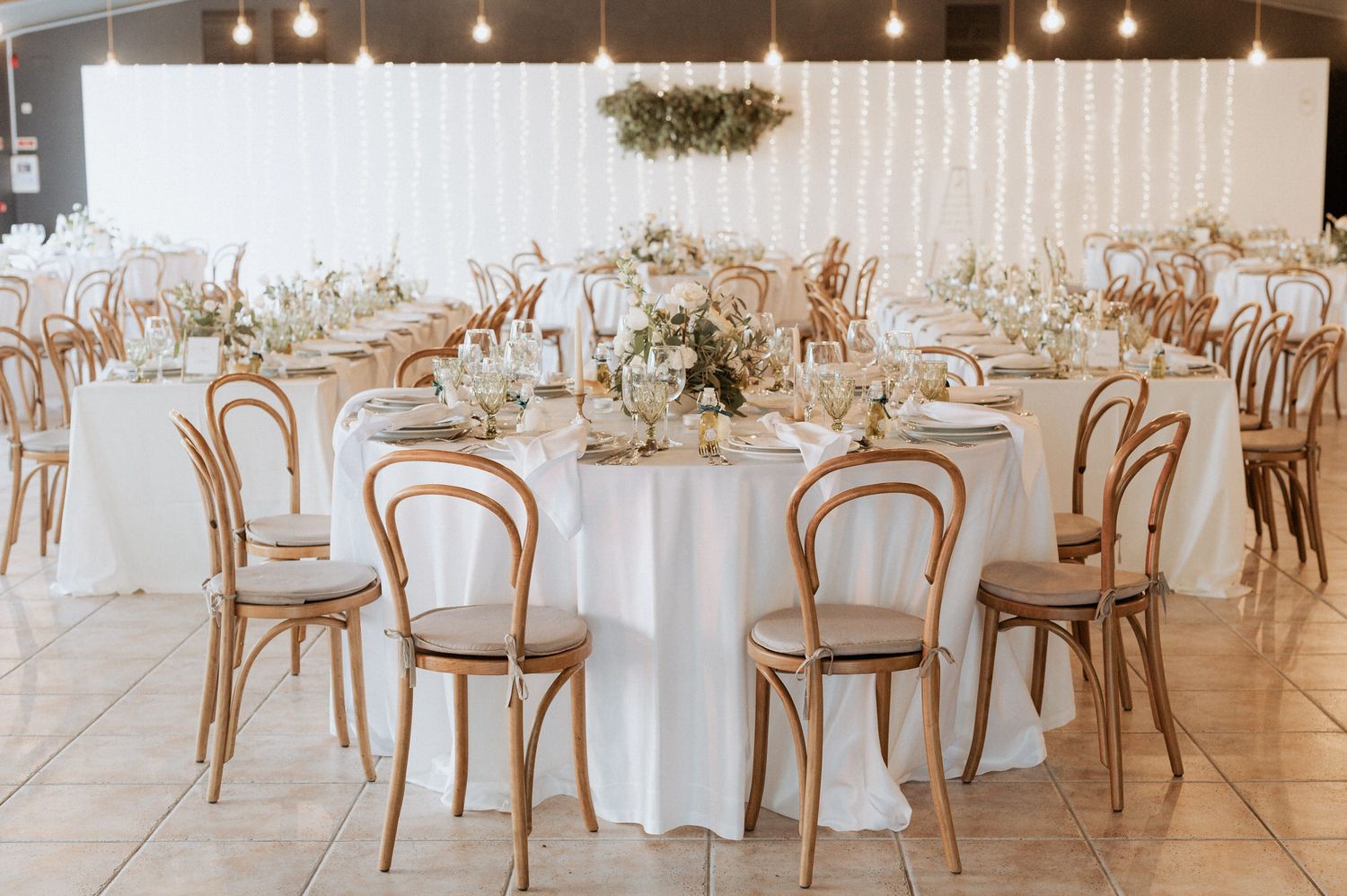 Elegant wedding reception venue with white round tables, wooden Bentwood chairs, and pendant lighting creating a romantic atmosphere.