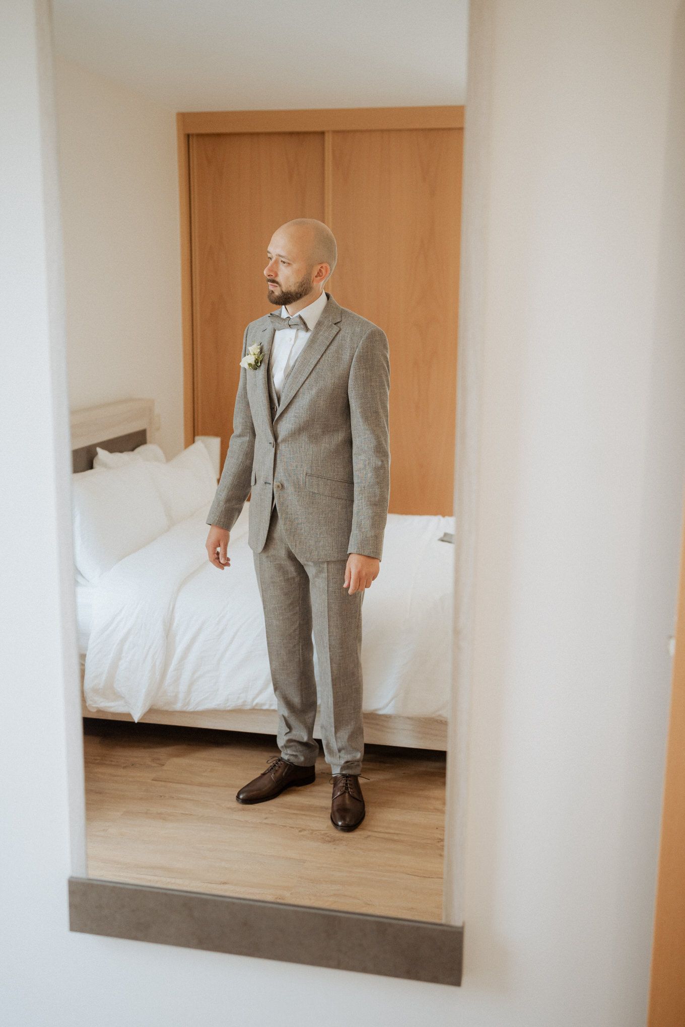 A groom in a light gray suit and bow tie stands in a minimalist hotel room with white bedding and wooden floors.