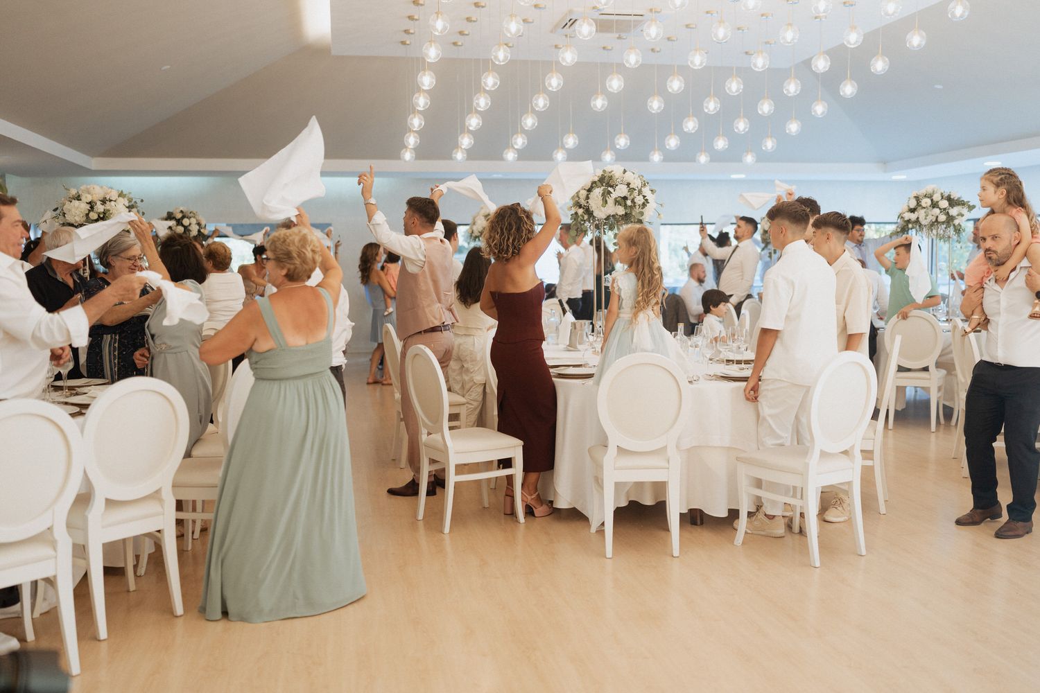 Wedding reception guests standing and celebrating at an elegant white-themed venue with sparkling overhead lights.