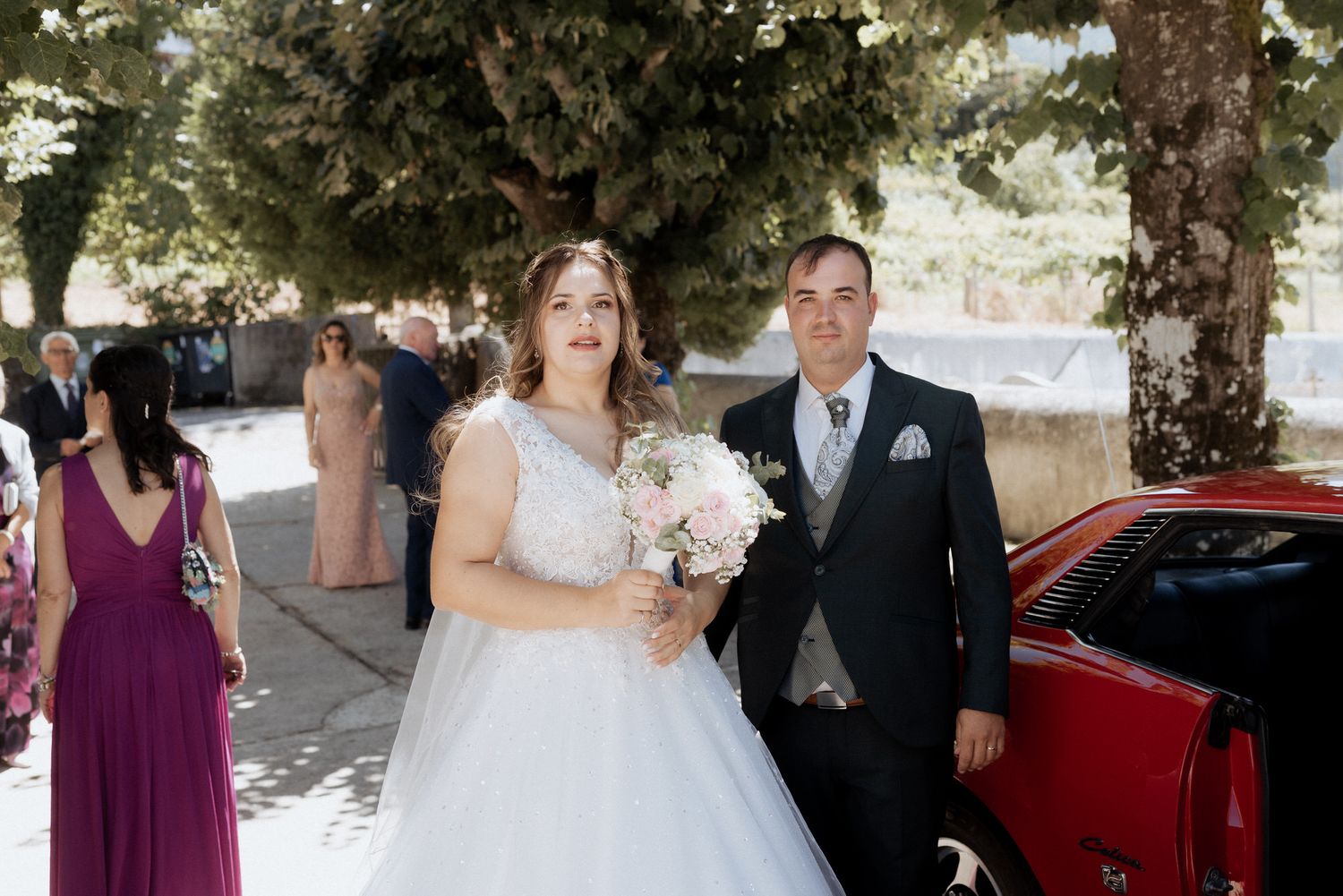 A bride in a white lace gown holding a bouquet walks beside someone in a dark suit on a tree-lined path.
