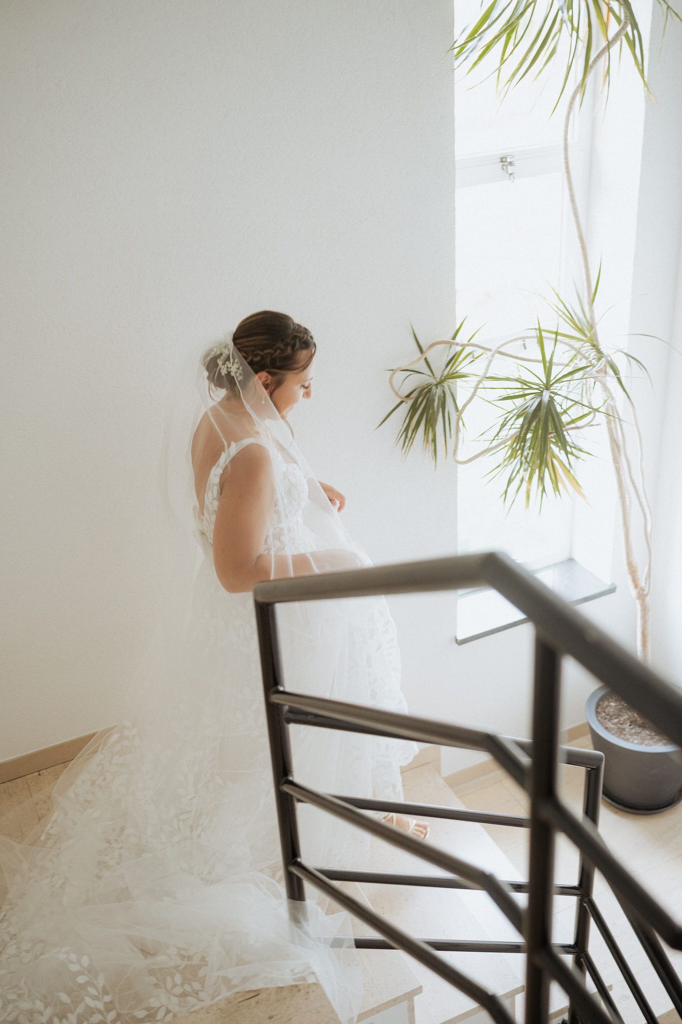 A bride in a flowing white wedding dress stands by a modern staircase near a sunlit window with tropical plants.