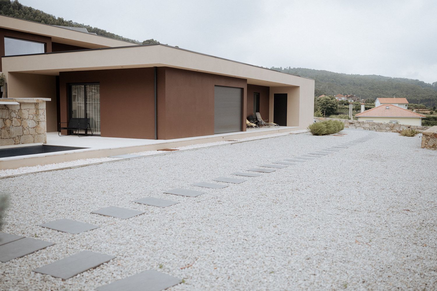 Modern brown minimalist house with gravel driveway and mountain views in the background.