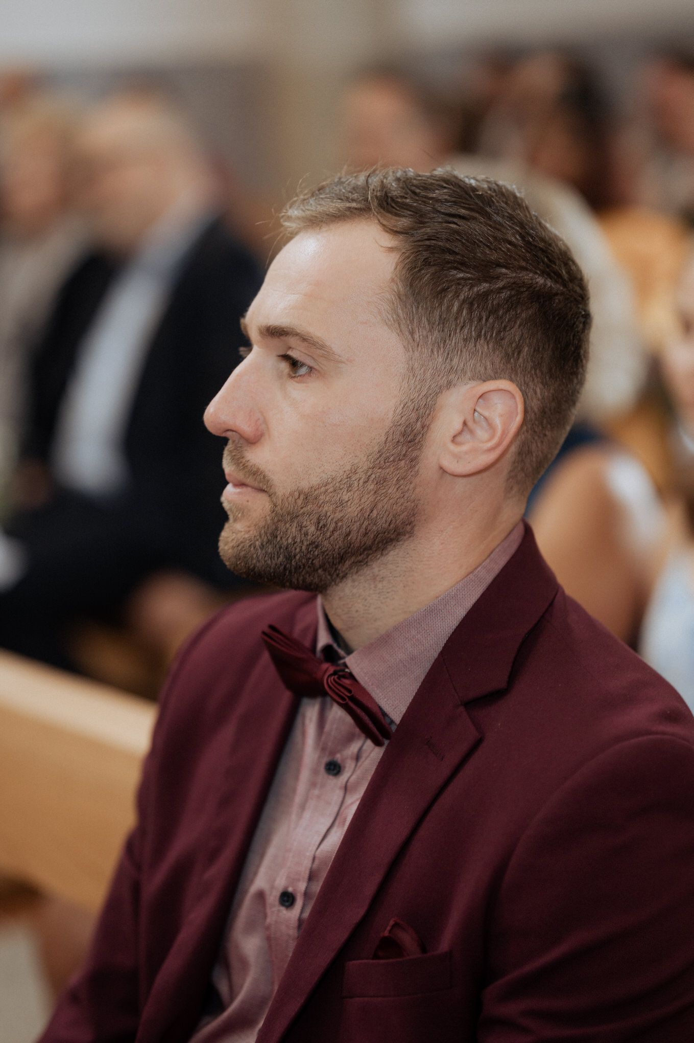 A person in a burgundy blazer and bow tie sits attentively in a formal setting.