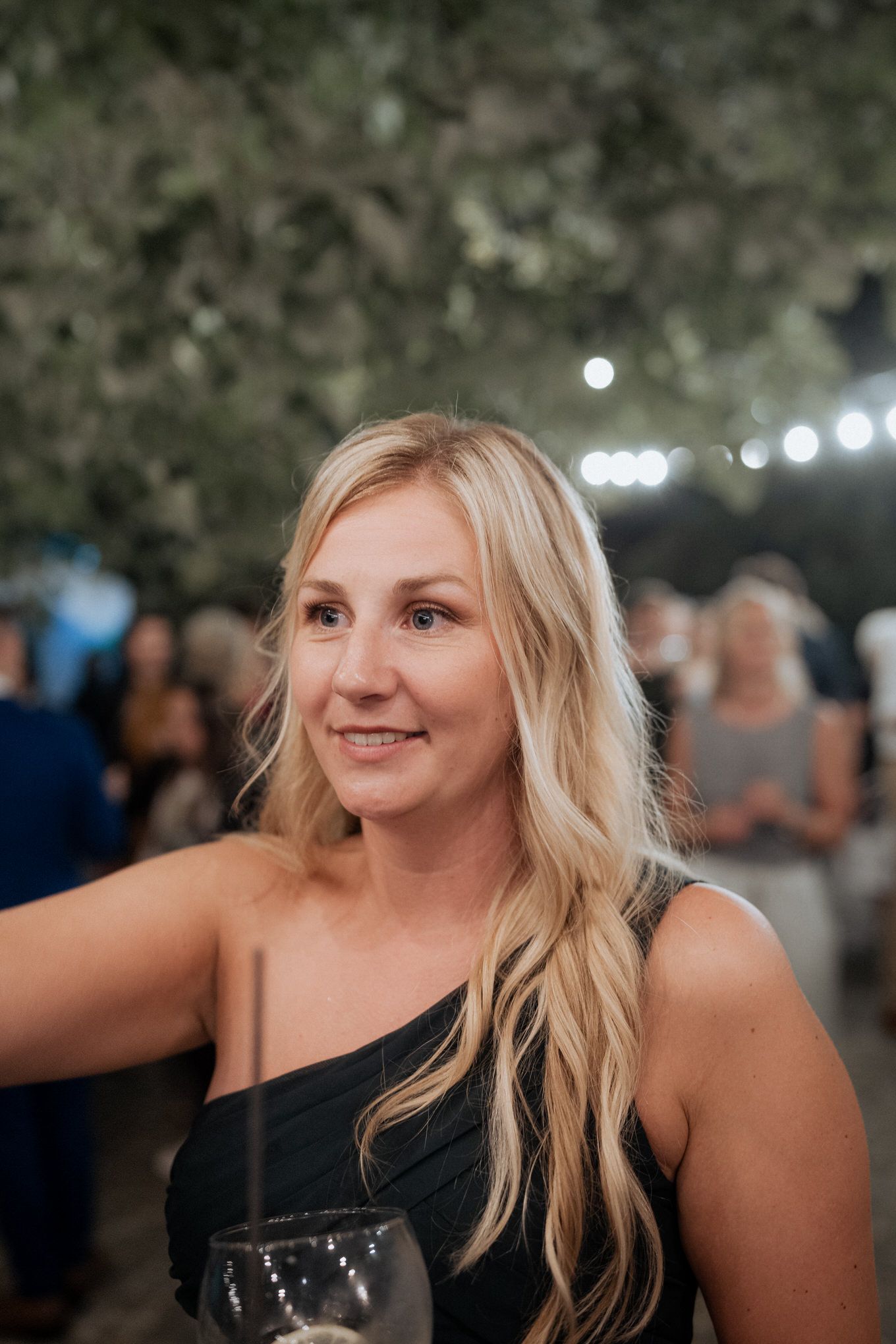 Woman with long blonde hair wearing a black one-shoulder top smiles at outdoor event with string lights in background.