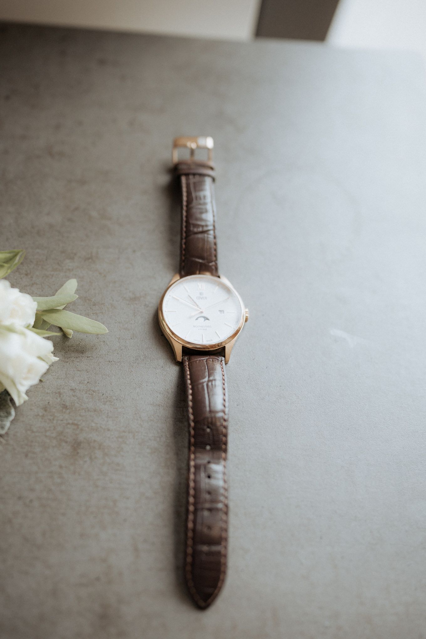 Classic gold watch with brown leather band on gray surface next to white flower.