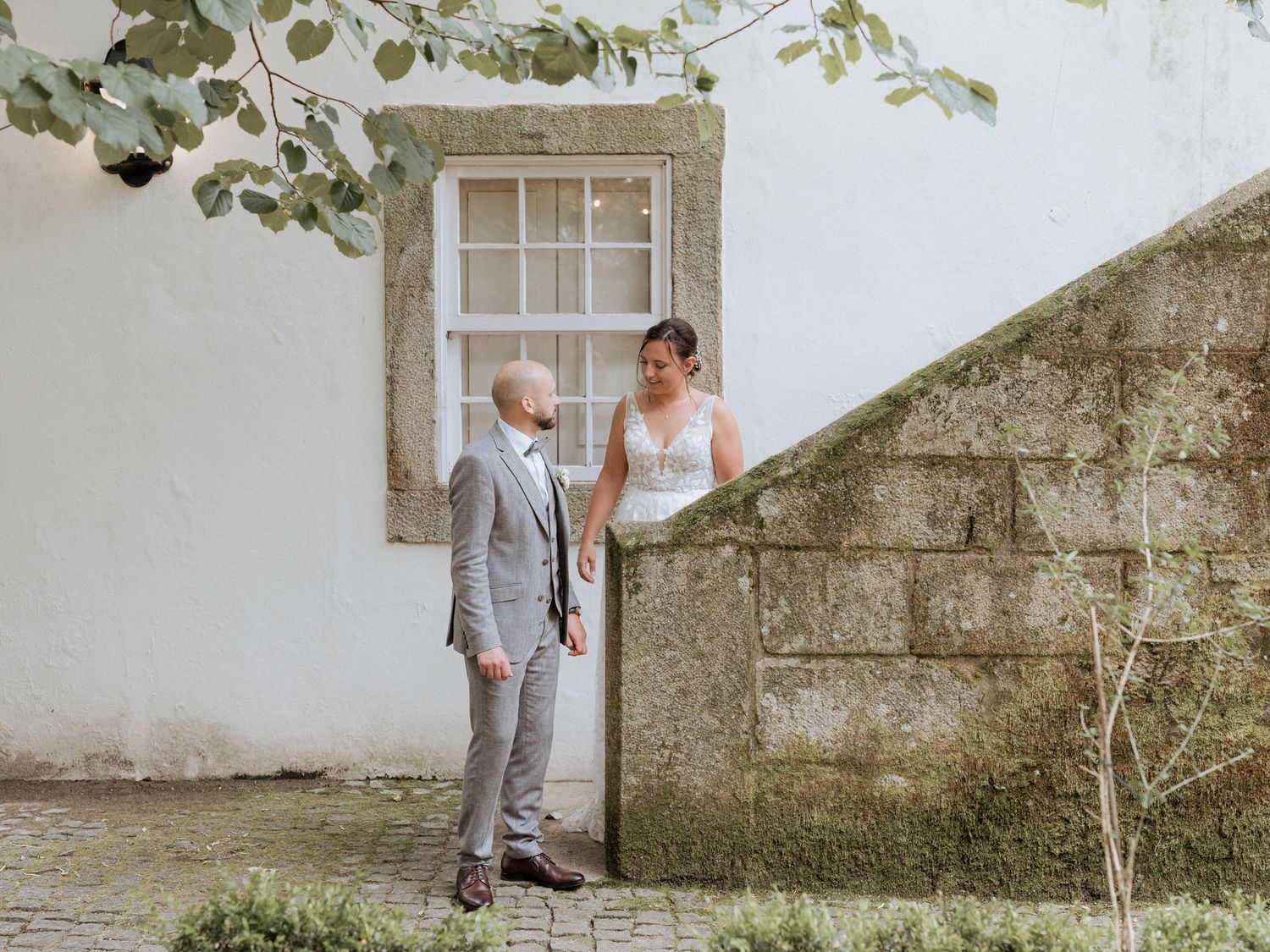 A couple shares a romantic moment outdoors by a white wall with vintage window frame and stone elements.