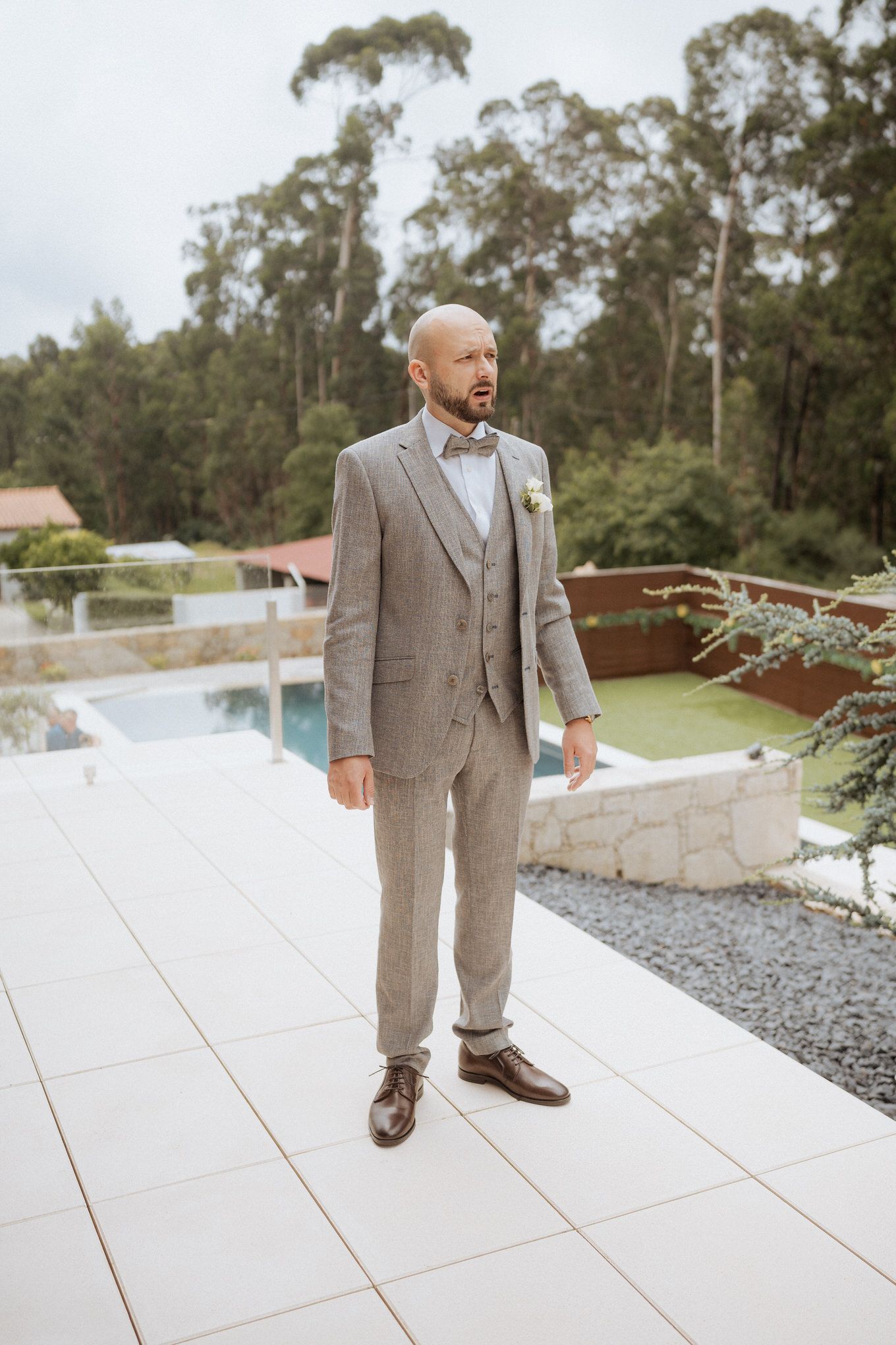 Groom in a tailored gray suit with a bow tie stands on a white tiled patio with trees in the background.