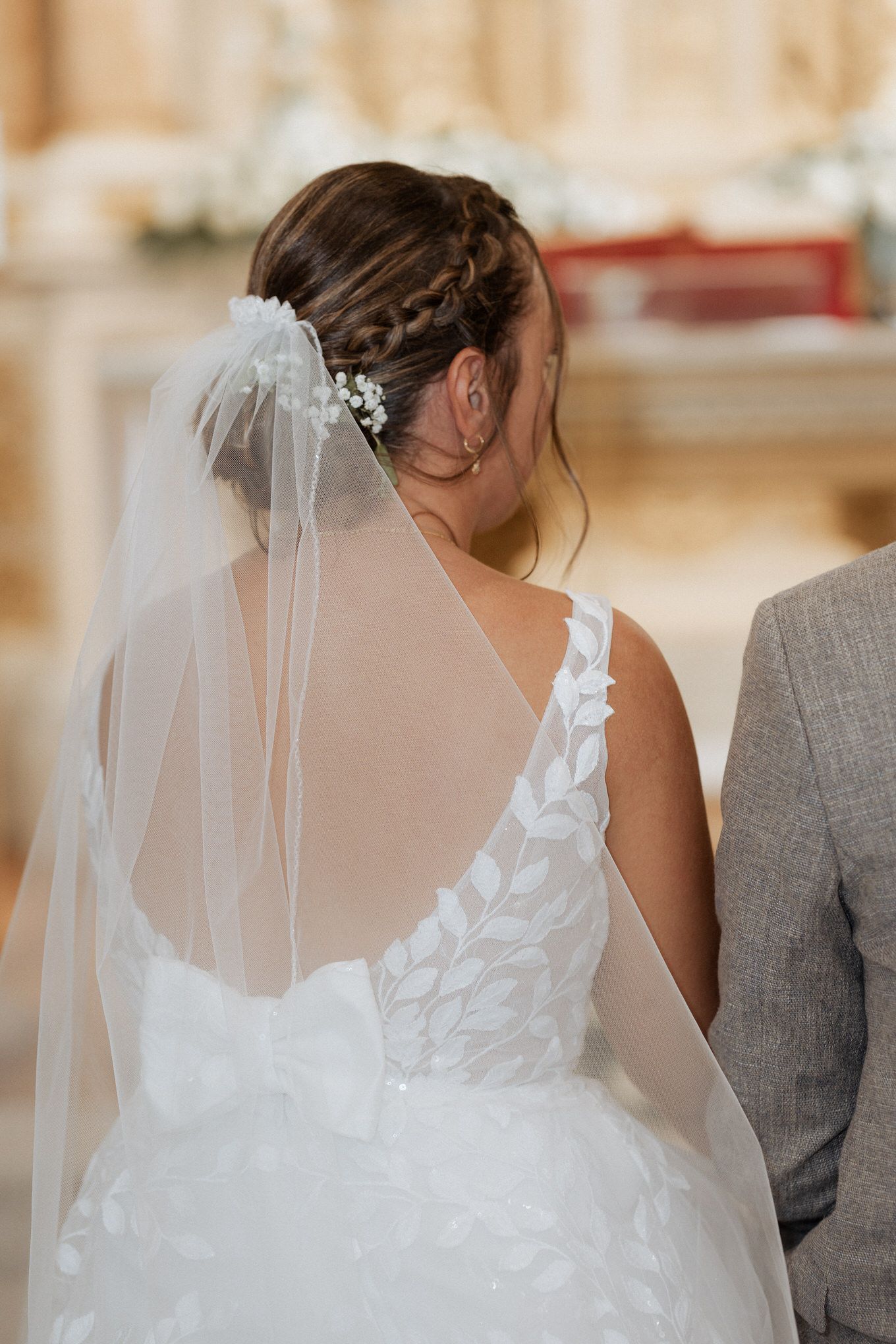 A bride in a white lace wedding dress with a flowing veil stands beside someone in gray during a wedding ceremony.