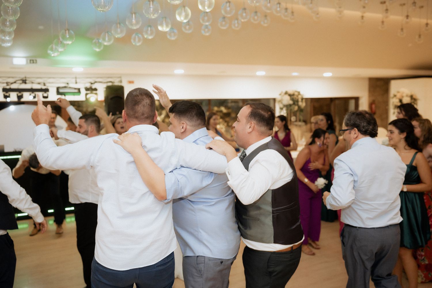 People dancing together in a conga line at a celebratory reception with overhead lighting and decorative elements.