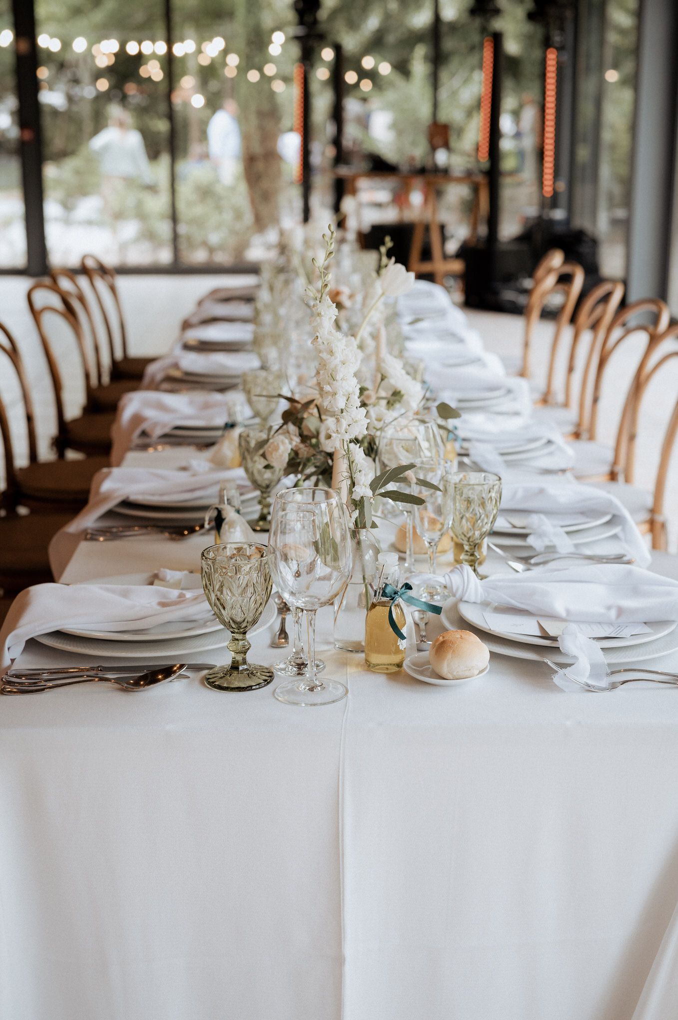 Elegant wedding table setting with white floral centerpiece, vintage glassware, and bentwood chairs in a sun-lit venue.