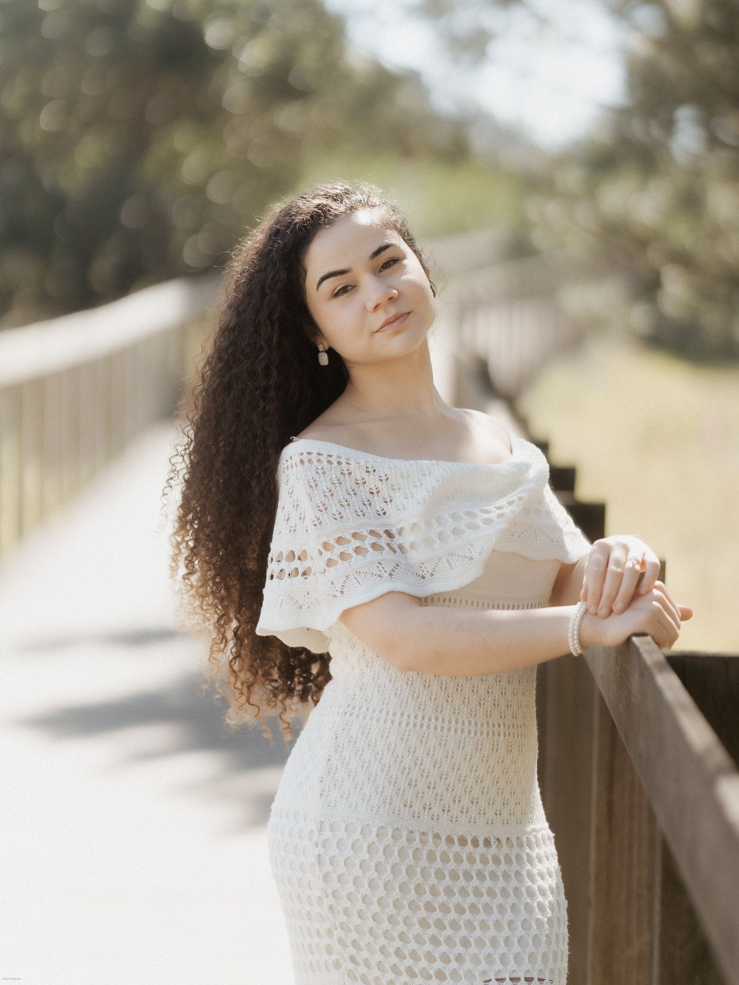 Jovem mulher com vestido de crochê branco posa em uma ponte de madeira durante um dia ensolarado com cenário natural ao ar livre.