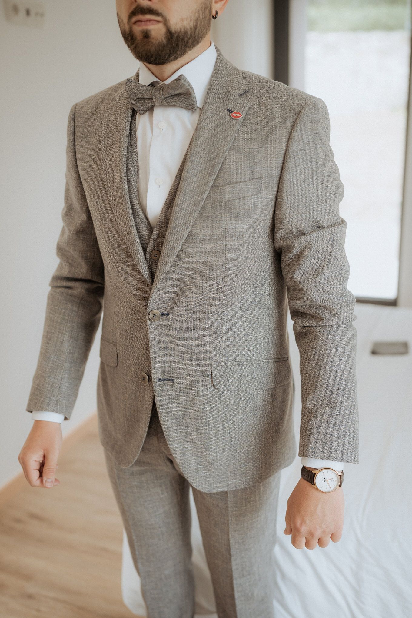 A stylish light gray three-piece suit with bow tie and red lapel pin, showcasing formal wedding attire details.