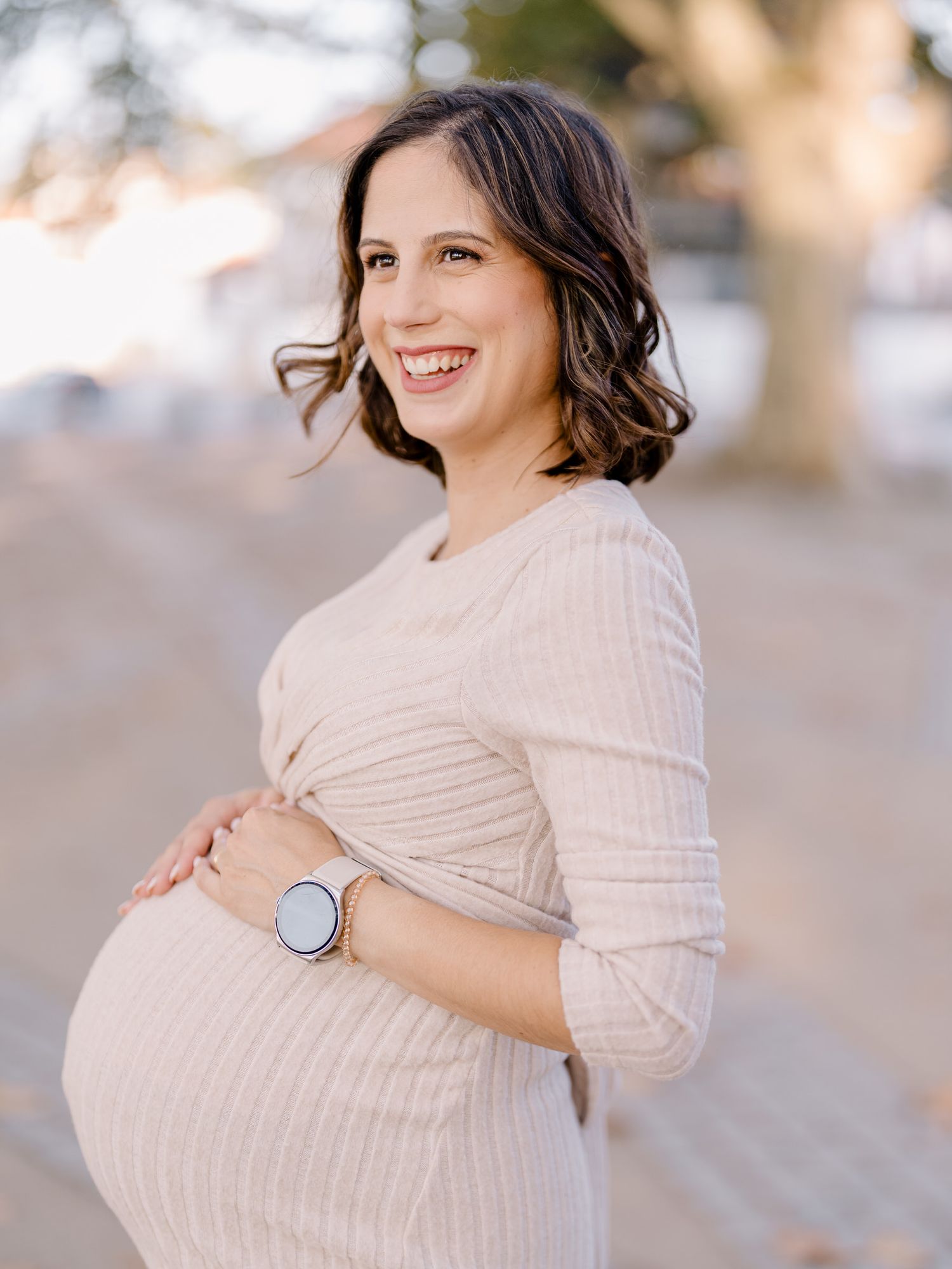 Uma futura mamãe usando um vestido de malha bege está ao ar livre sorrindo enquanto segura sua barriga de grávida e usa um relógio prateado.