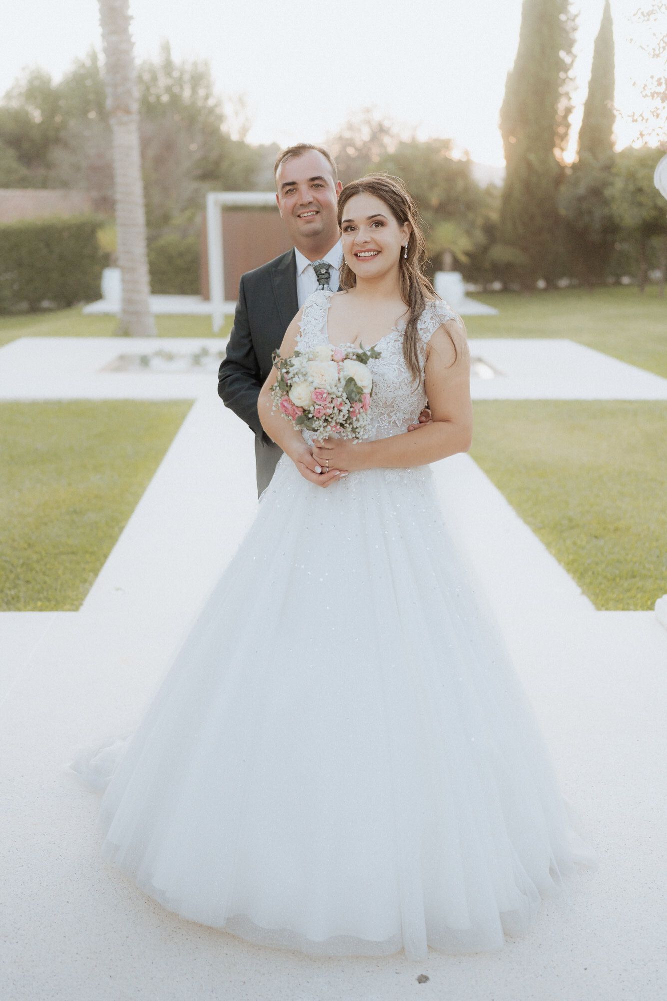A joyful couple poses outdoors at golden hour, with the bride wearing a full white ballgown and holding a pink bouquet.