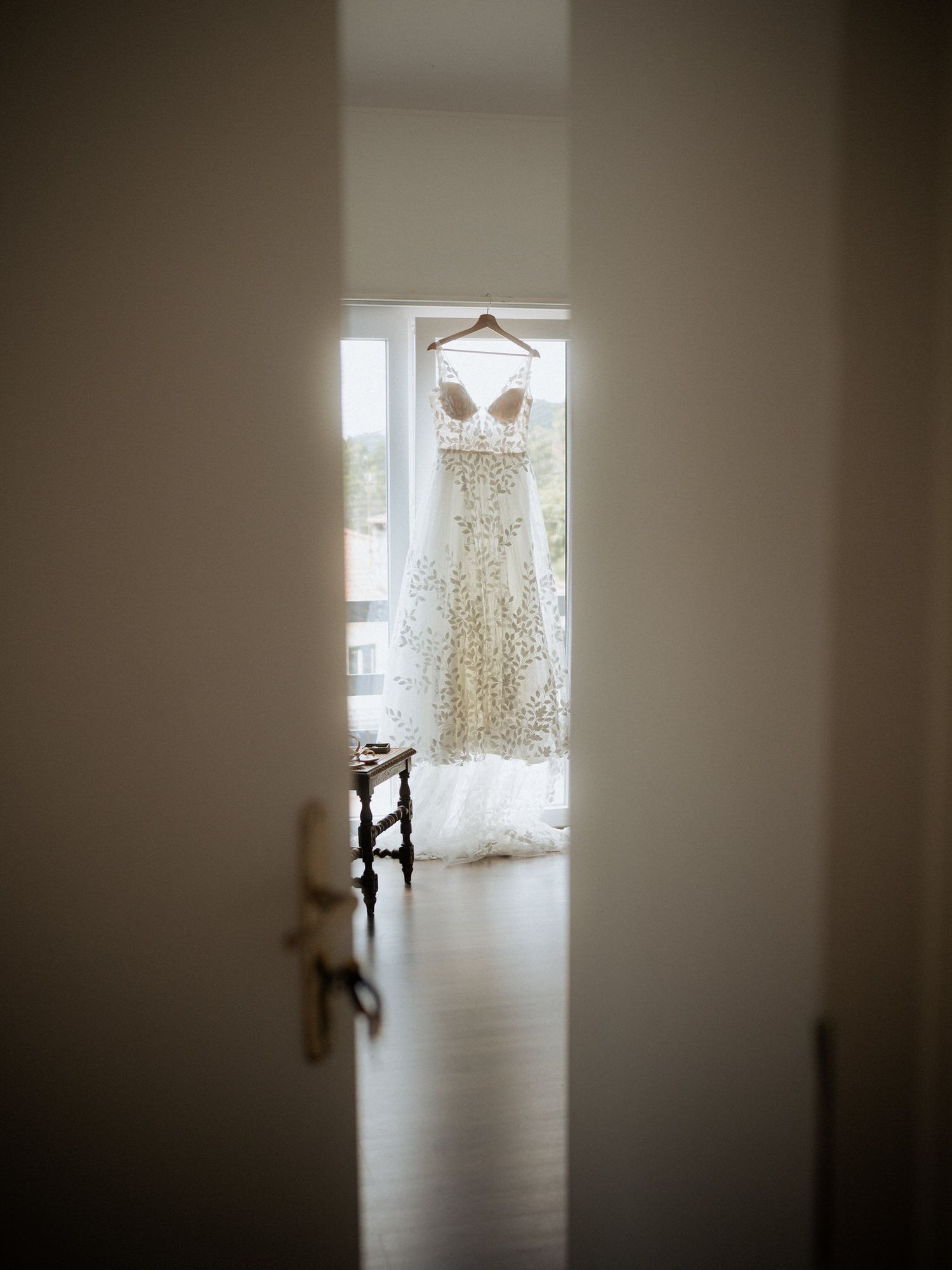 A delicate lace wedding dress hangs in a sunlit doorway, photographed through an open interior door.