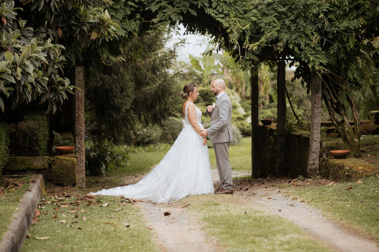 A couple shares a romantic moment on a tree-lined garden path on their wedding day.
