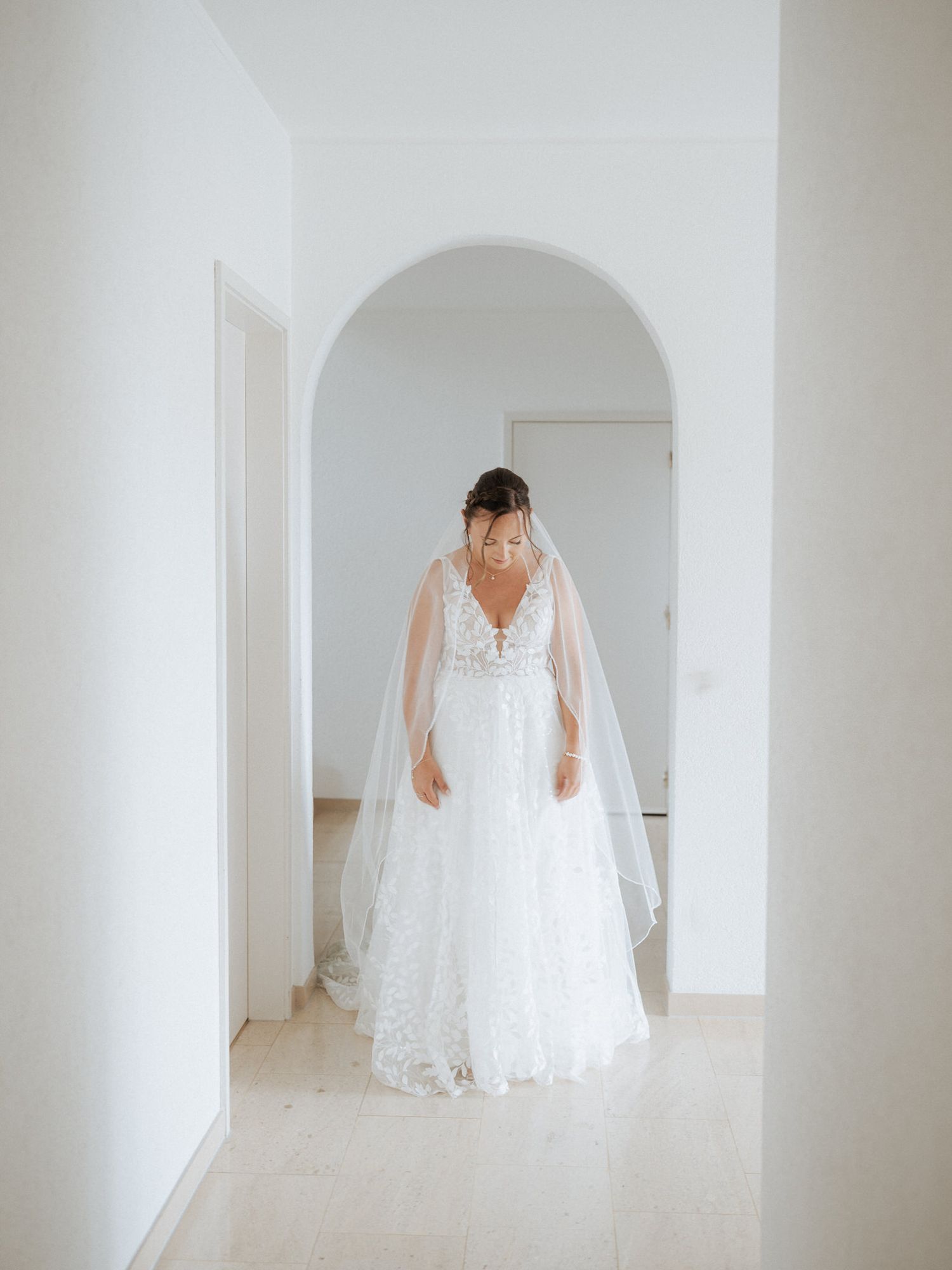 An elegant white lace wedding gown with a deep V neckline and flowing veil photographed in a minimalist white hallway.
