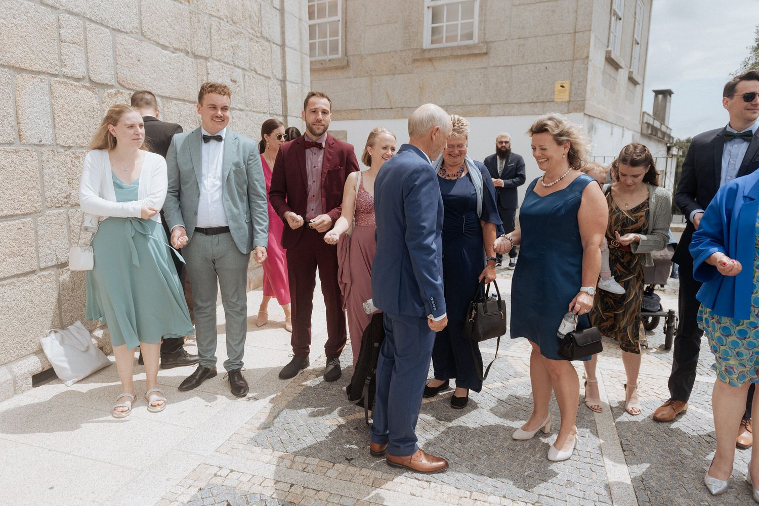 A group of elegantly dressed wedding guests gather outside a stone building on a sunny day.