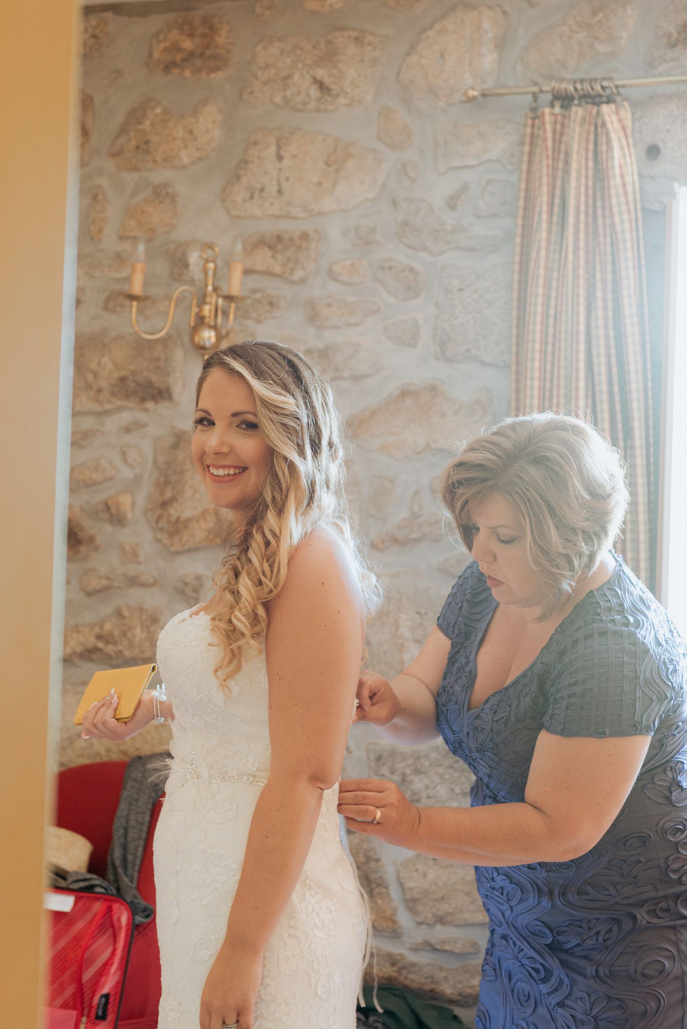 Series of photos showing a bride getting into her wedding dress with help from someone in a blue dress beside stone walls.