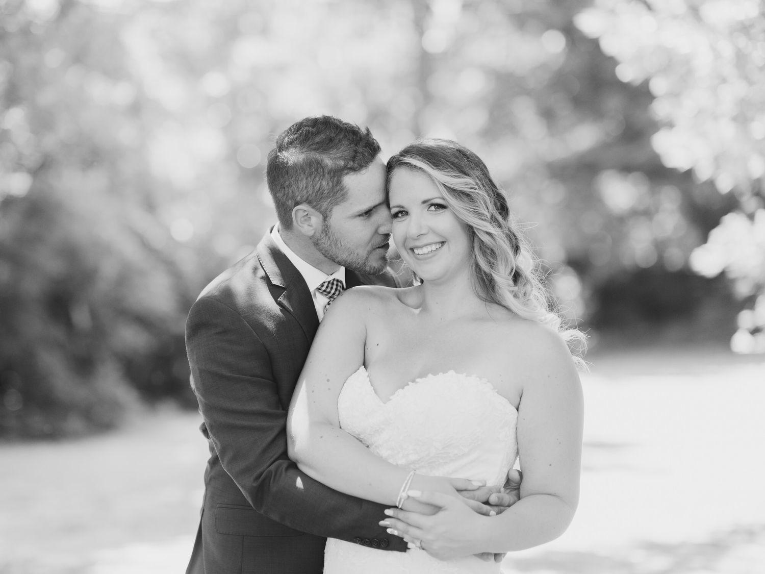 A couple embraces on their wedding day outdoors in black and white with soft bokeh lighting.