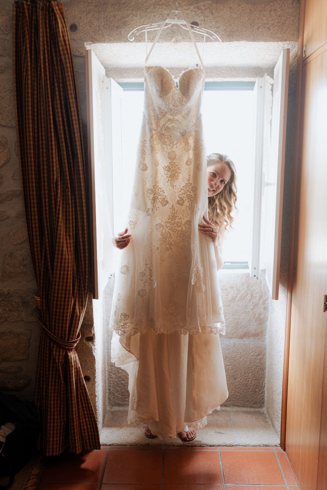 A delicate white lace wedding dress hangs in front of a bright window with someone peeking out from behind.