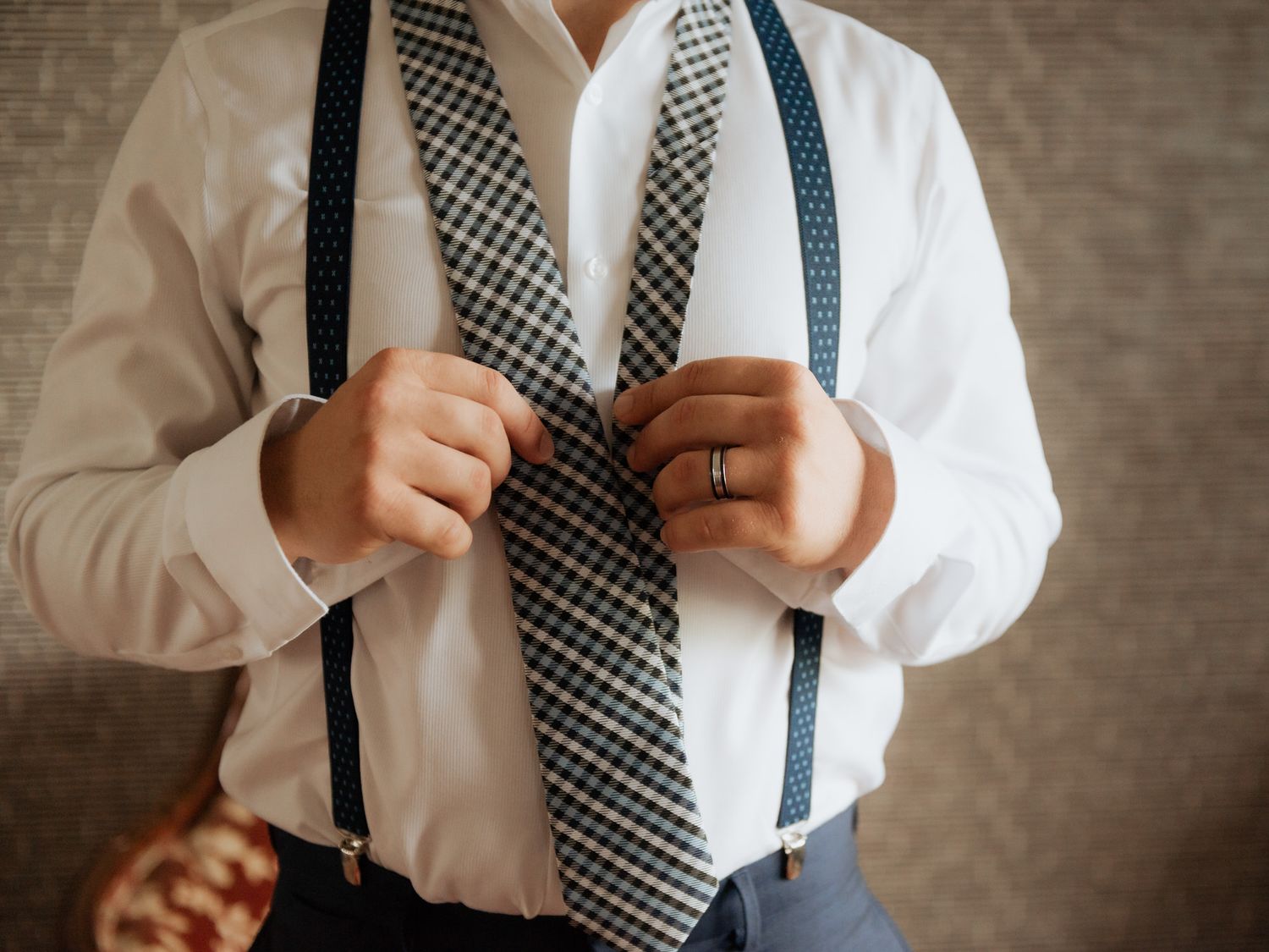 Person wearing a white dress shirt adjusts navy blue suspenders and checkered tie against textured wall.